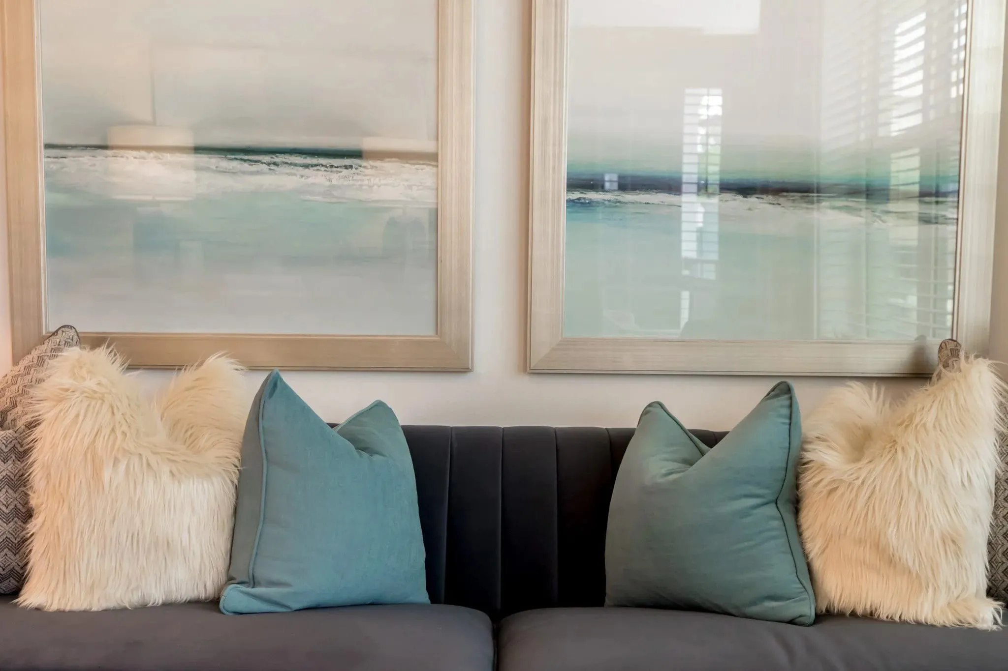Multifamily interior detail: charcoal channel-back sofa with teal and faux fur pillows below two coastal paintings
