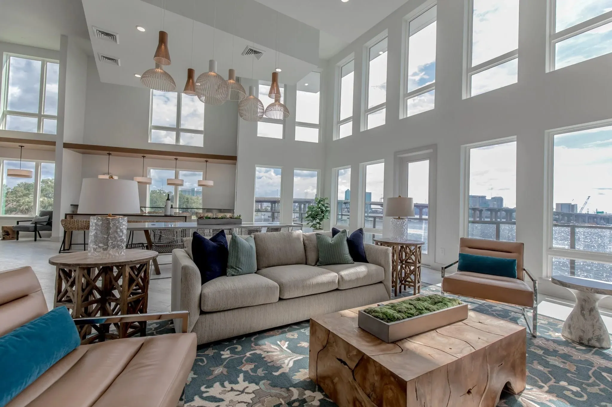 Multifamily amenity clubroom lounge with sofas and a live edge coffee table below woven pendants beside the river windows