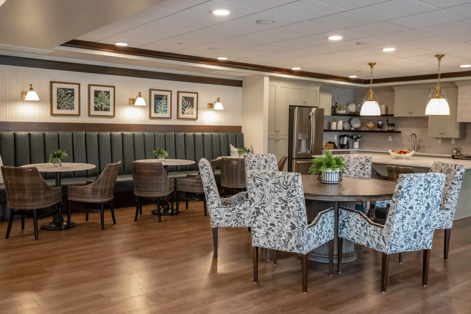 Senior living bistro with a dark green banquette under botanical prints, rattan and floral dining chairs by an open kitchen