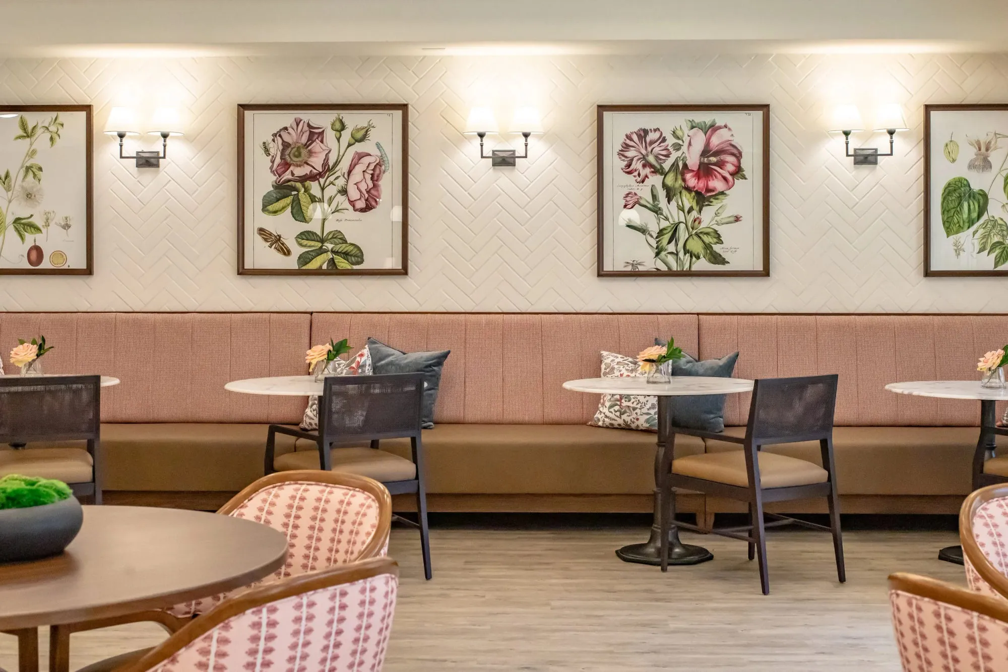 Senior living bistro with a blush upholstered banquette, round cafe tables and framed botanical prints on white herringbone tile