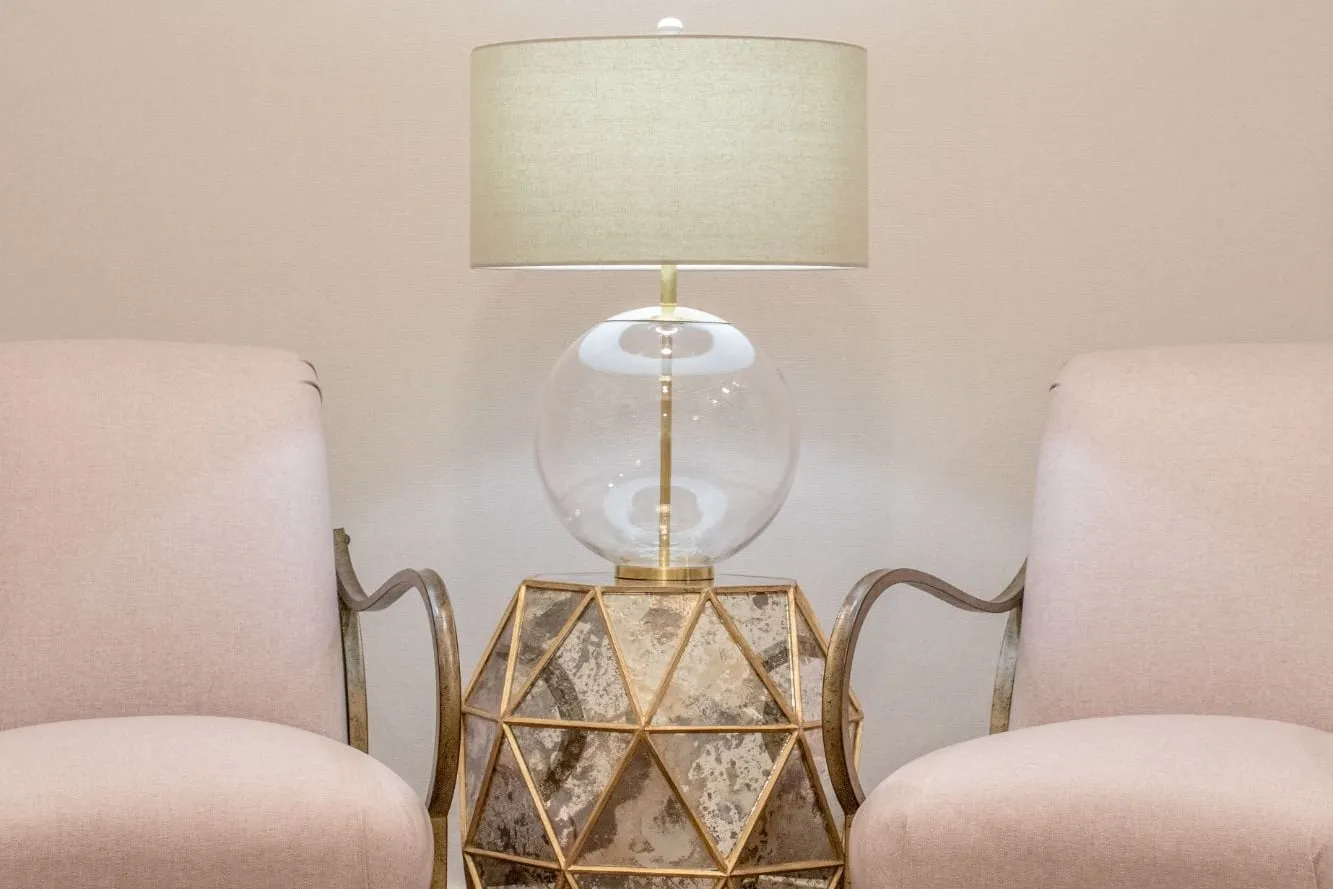 Senior living sitting area with two blush armchairs on curved metal arms either side of a gold faceted antique mirror side table and a clear glass globe lamp