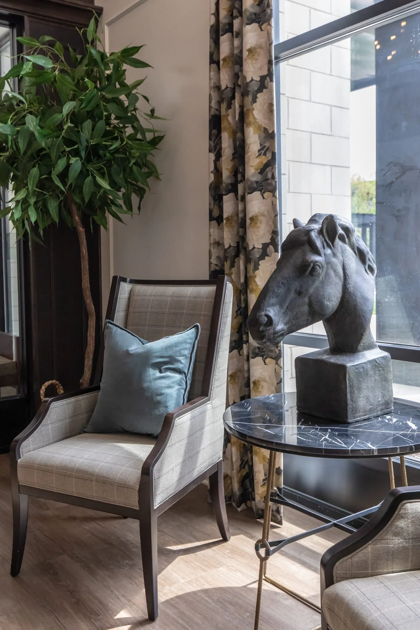 Grey armchair with a blue pillow beside a bronze horse head sculpture on a side table, in front of tall windows