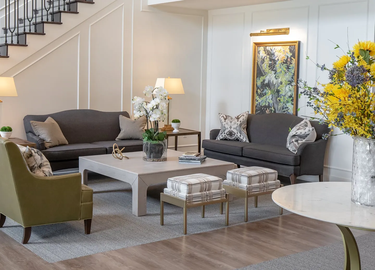 Senior living lounge with white panelled walls, an olive leather armchair, charcoal sofas, plaid upholstered benches and yellow flowers on a marble table
