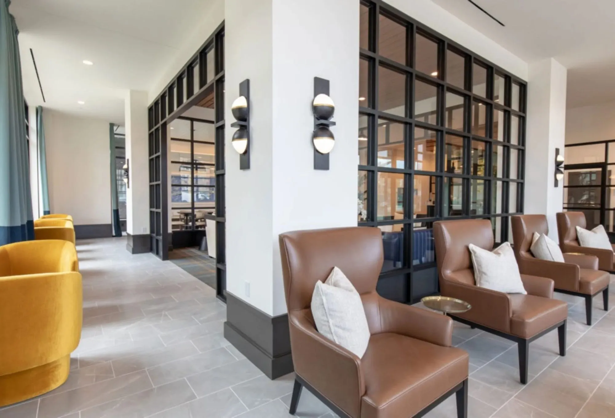 Senior living corridor lined with caramel leather wing chairs beside black steel and glass partitions, paired sphere sconces