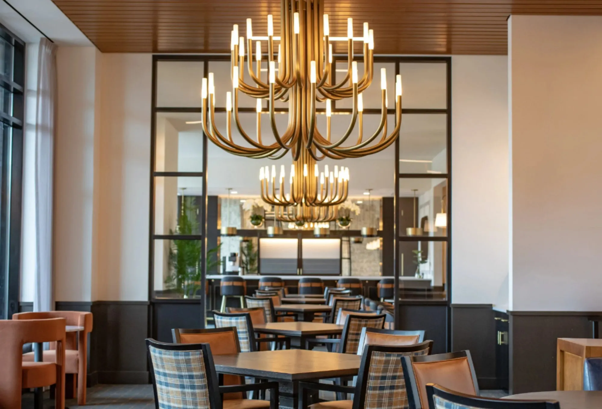 Senior living dining room with two brass candelabra chandeliers under a wood plank ceiling, plaid and caramel leather chairs