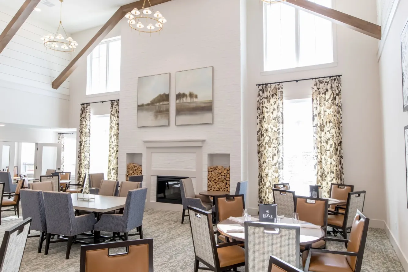 Senior living dining room under a vaulted ceiling with timber beams and brass ring chandeliers, a white brick fireplace between floral curtains, and cognac leather chairs at set tables