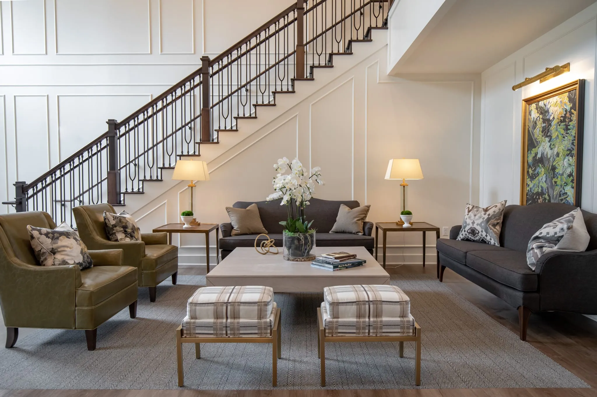 Two-storey lounge beneath an iron-balustrade staircase, with olive leather armchairs, charcoal sofas, plaid benches and white orchids on a square coffee table