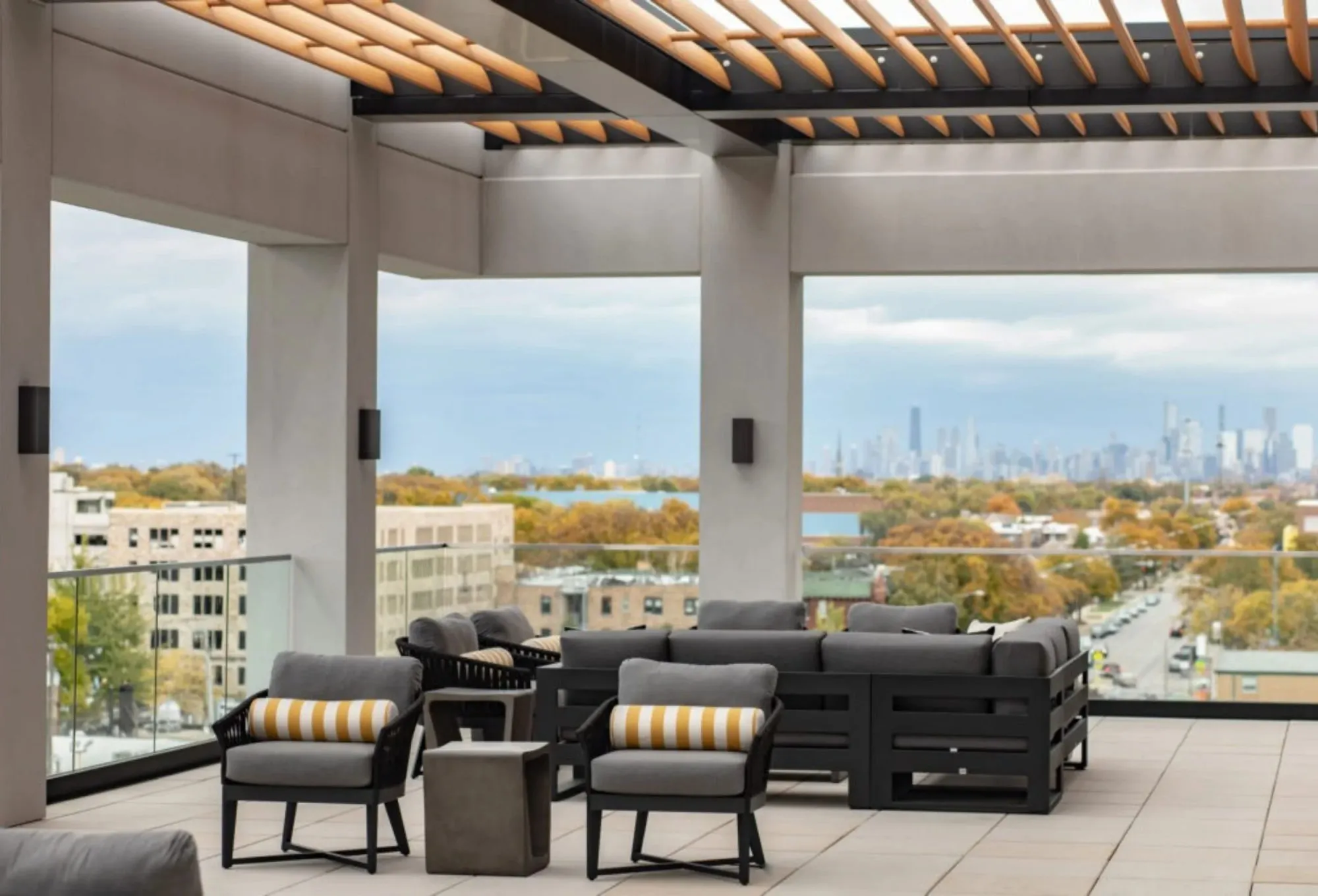 Senior living rooftop terrace under a wood slat pergola with a black sectional and yellow striped chairs, a skyline beyond