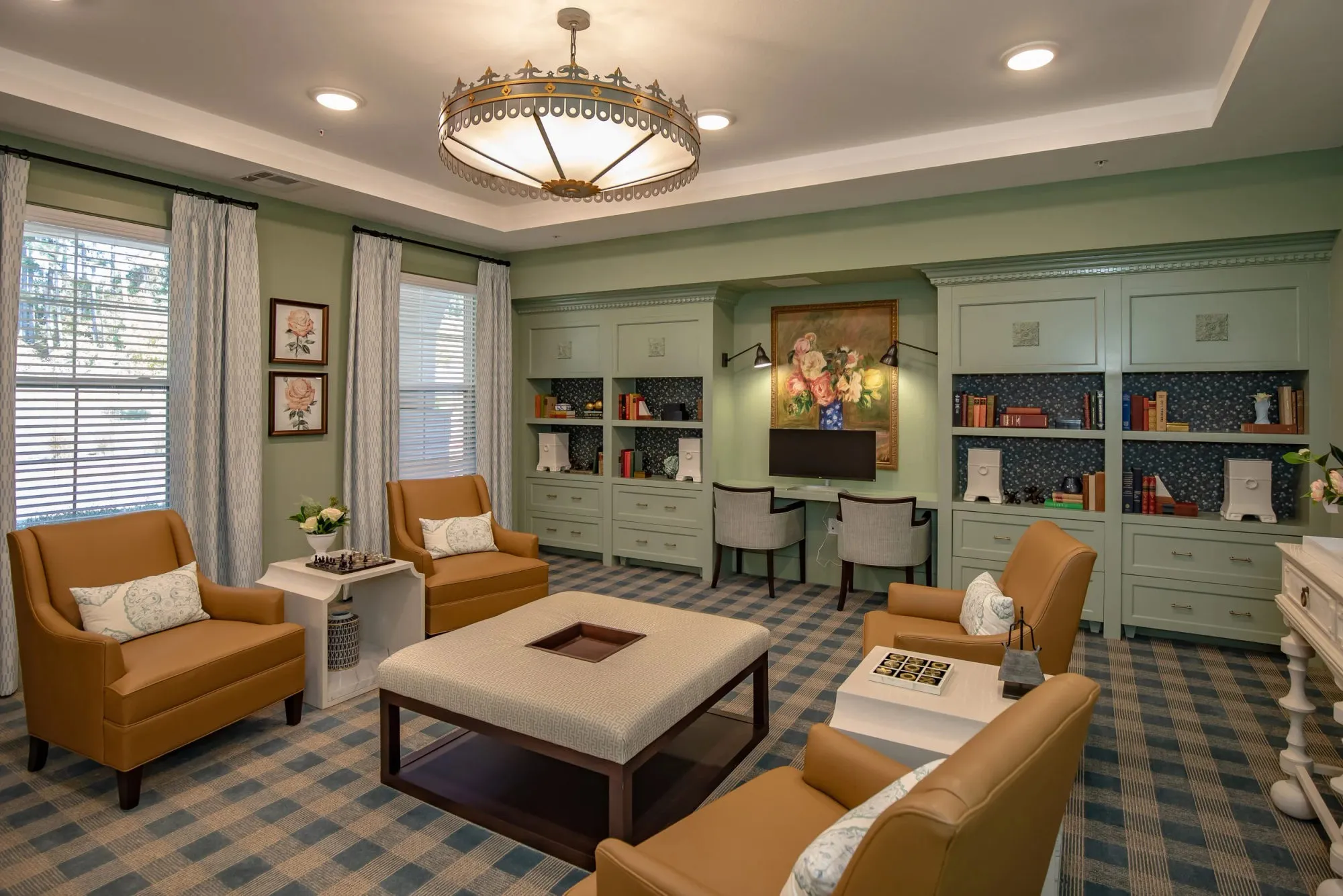 Senior living sitting room with sage green built-in cabinetry, camel armchairs, a fabric-topped table and blue plaid carpet