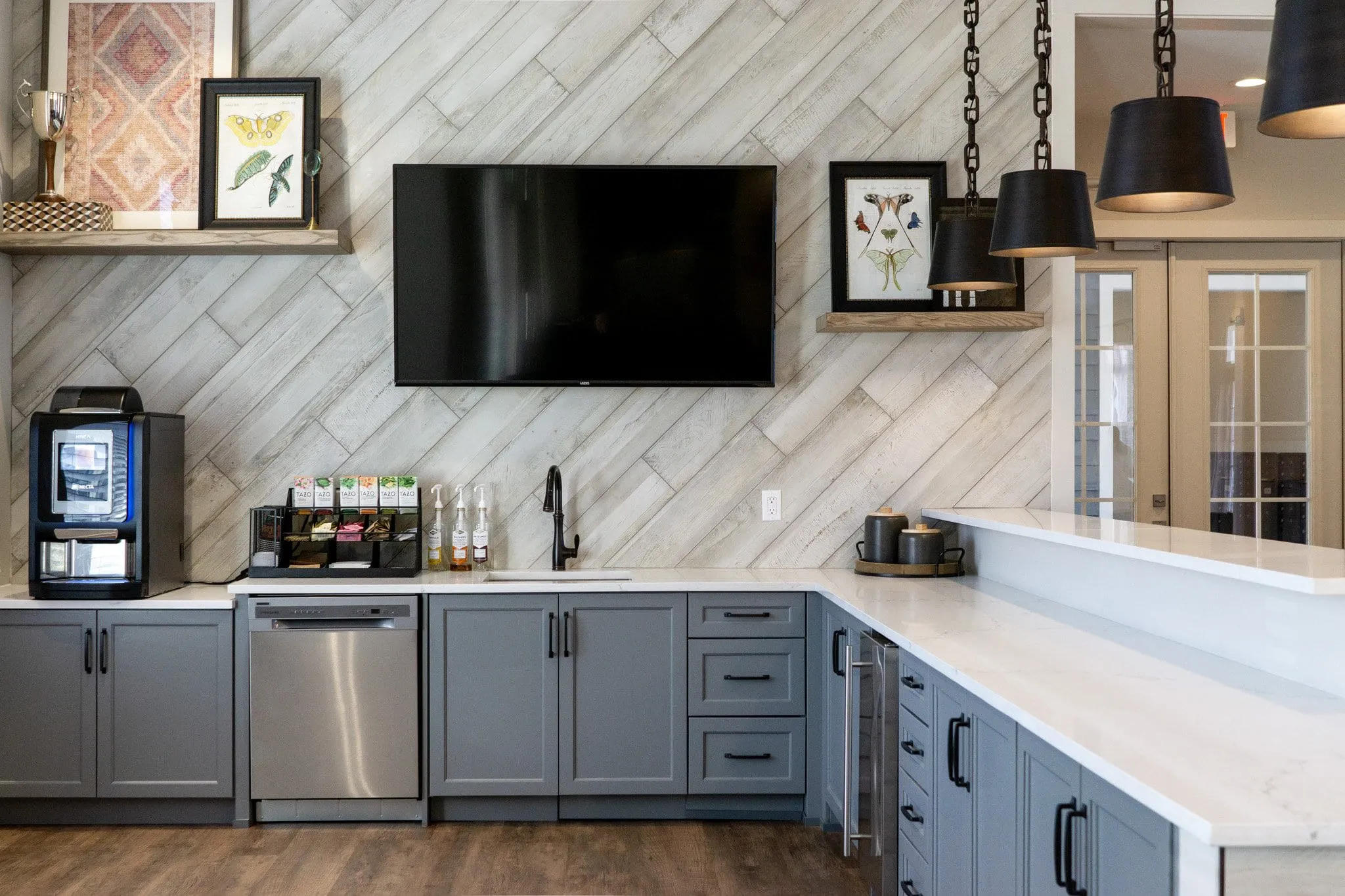 Multifamily amenity clubroom kitchen with grey cabinetry and white counters against a diagonal wood-look tile wall