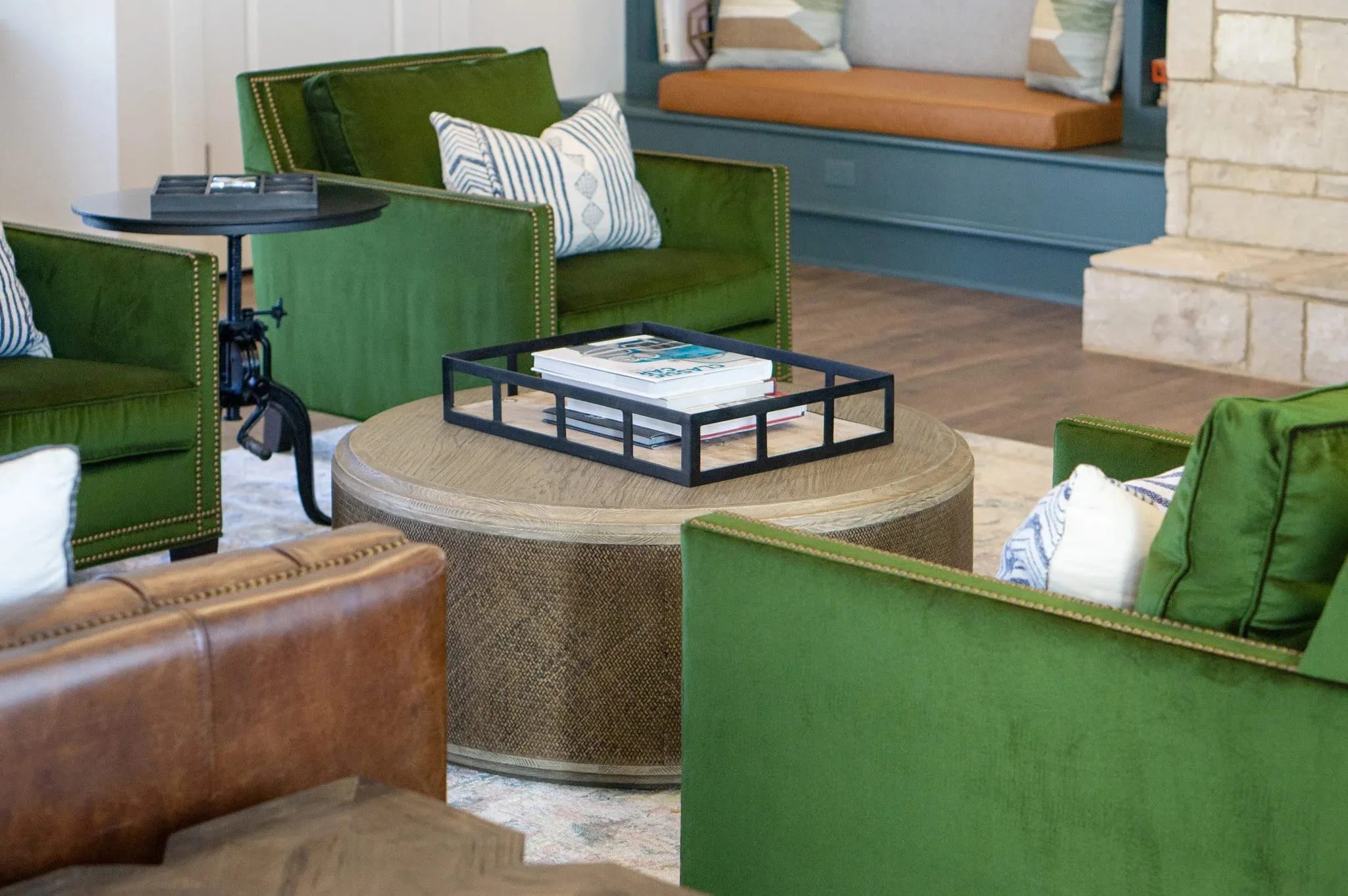 Multifamily amenity clubroom with green velvet chairs and a tufted leather sofa around a woven drum ottoman