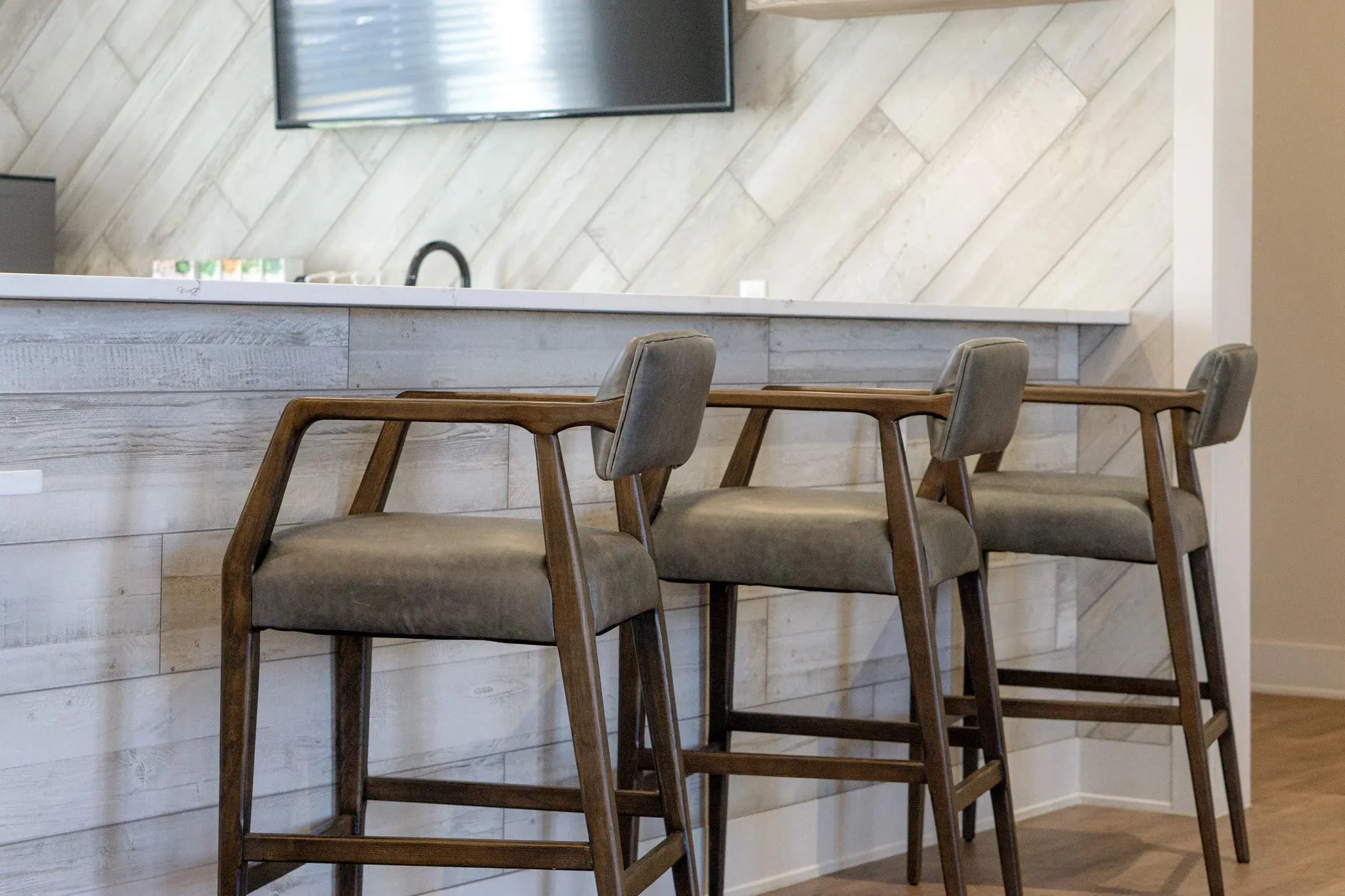 Multifamily amenity kitchen bar with three grey upholstered stools at a whitewashed plank counter, diagonal tile behind