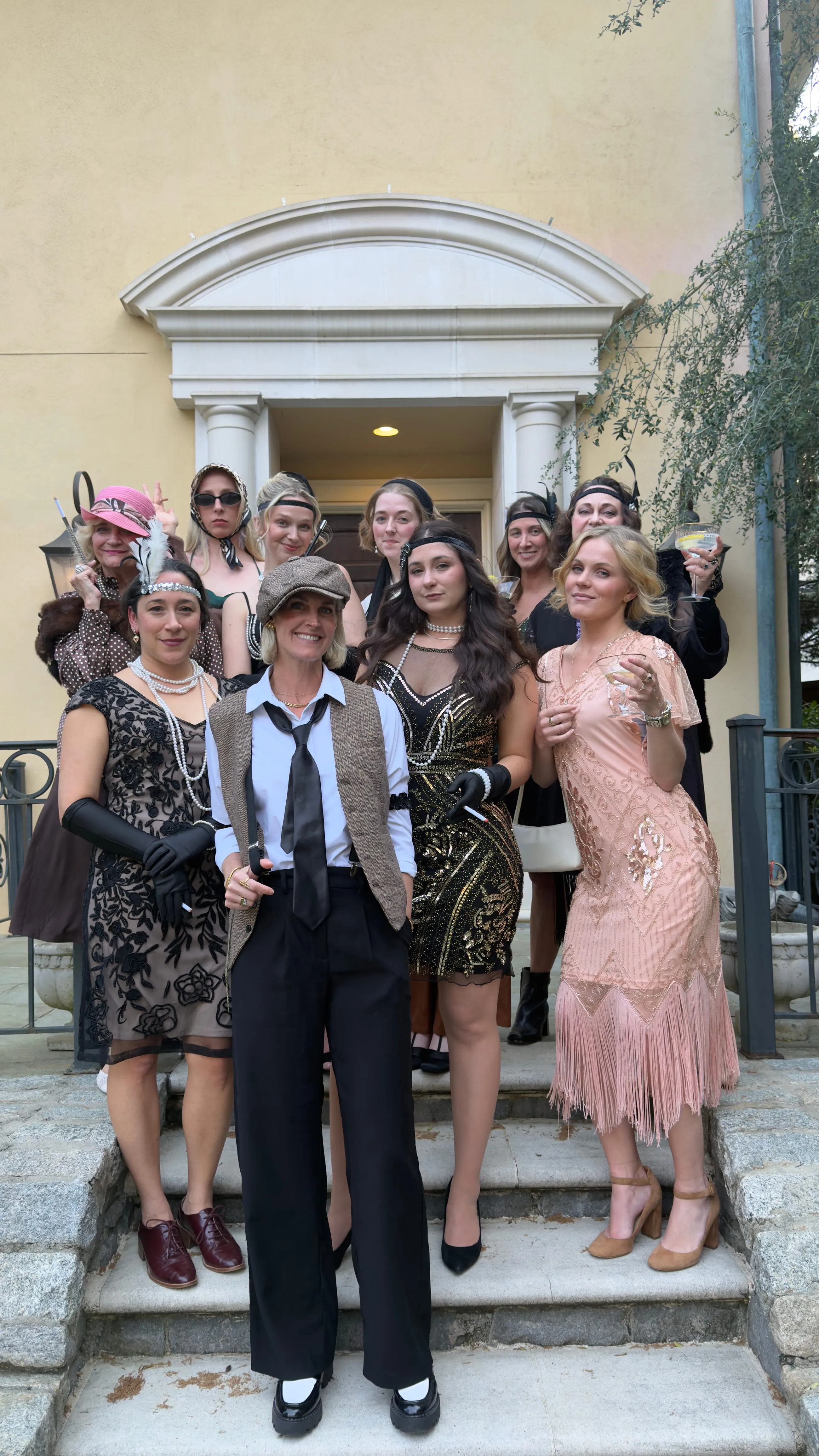 Five women in 1920s beaded dresses and headbands posing on the stone steps outside a house
