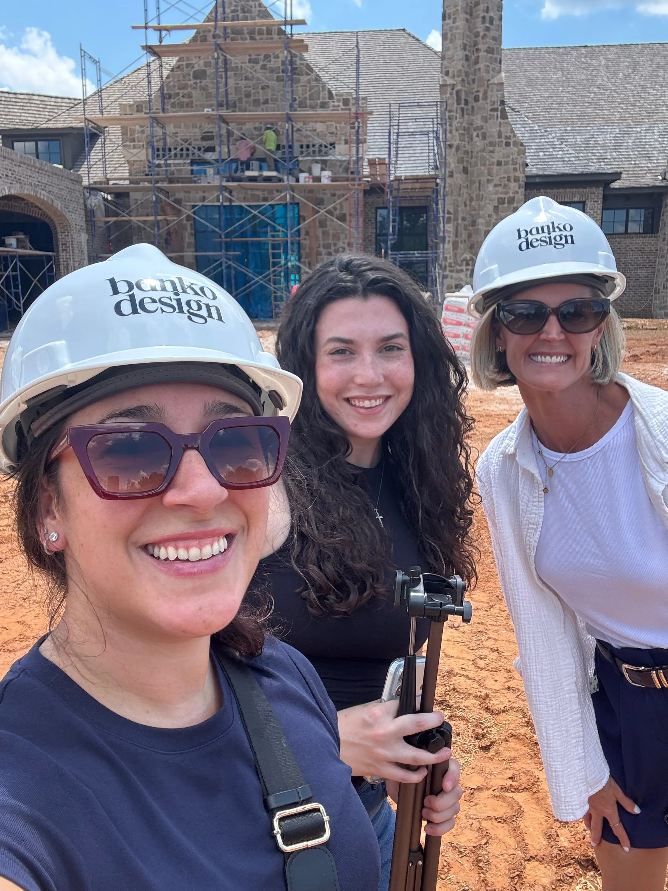 Three team members in white hard hats taking a selfie on a construction site, scaffolding on the building behind them