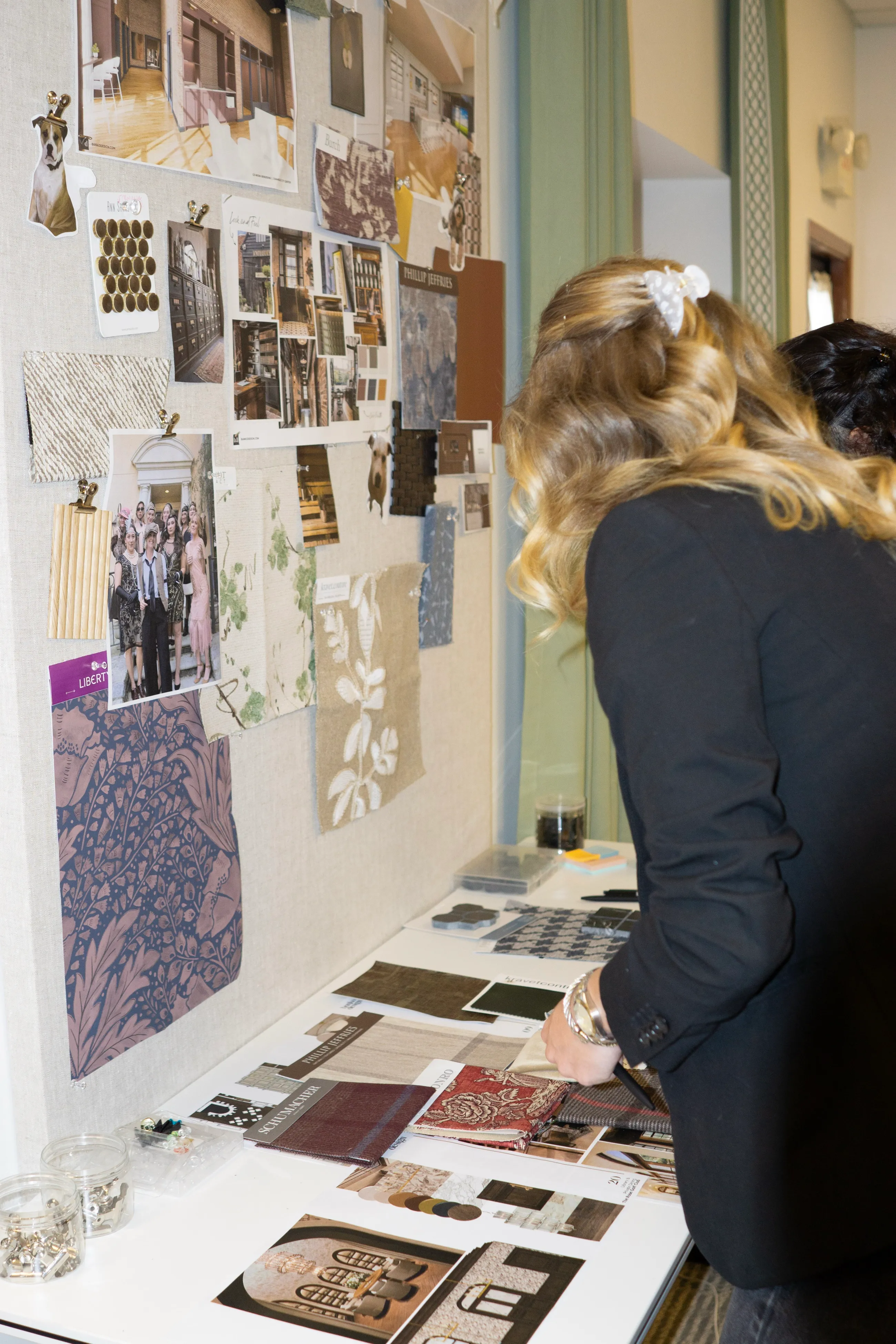 A Banko Design team member in a black blazer leaning over a table of fabric and material samples, beside a pin board of interior photographs and textiles