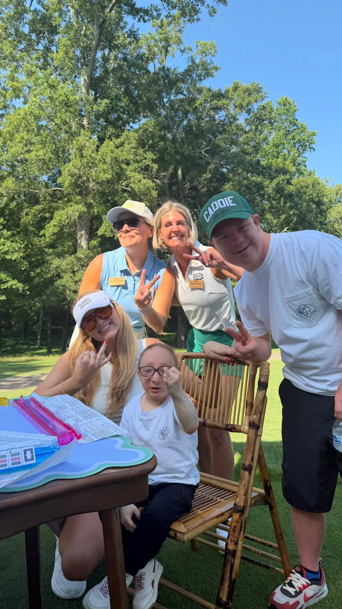 Five team members in caps and visors clowning for the camera around a folding table and a bamboo chair on a lawn under tall trees