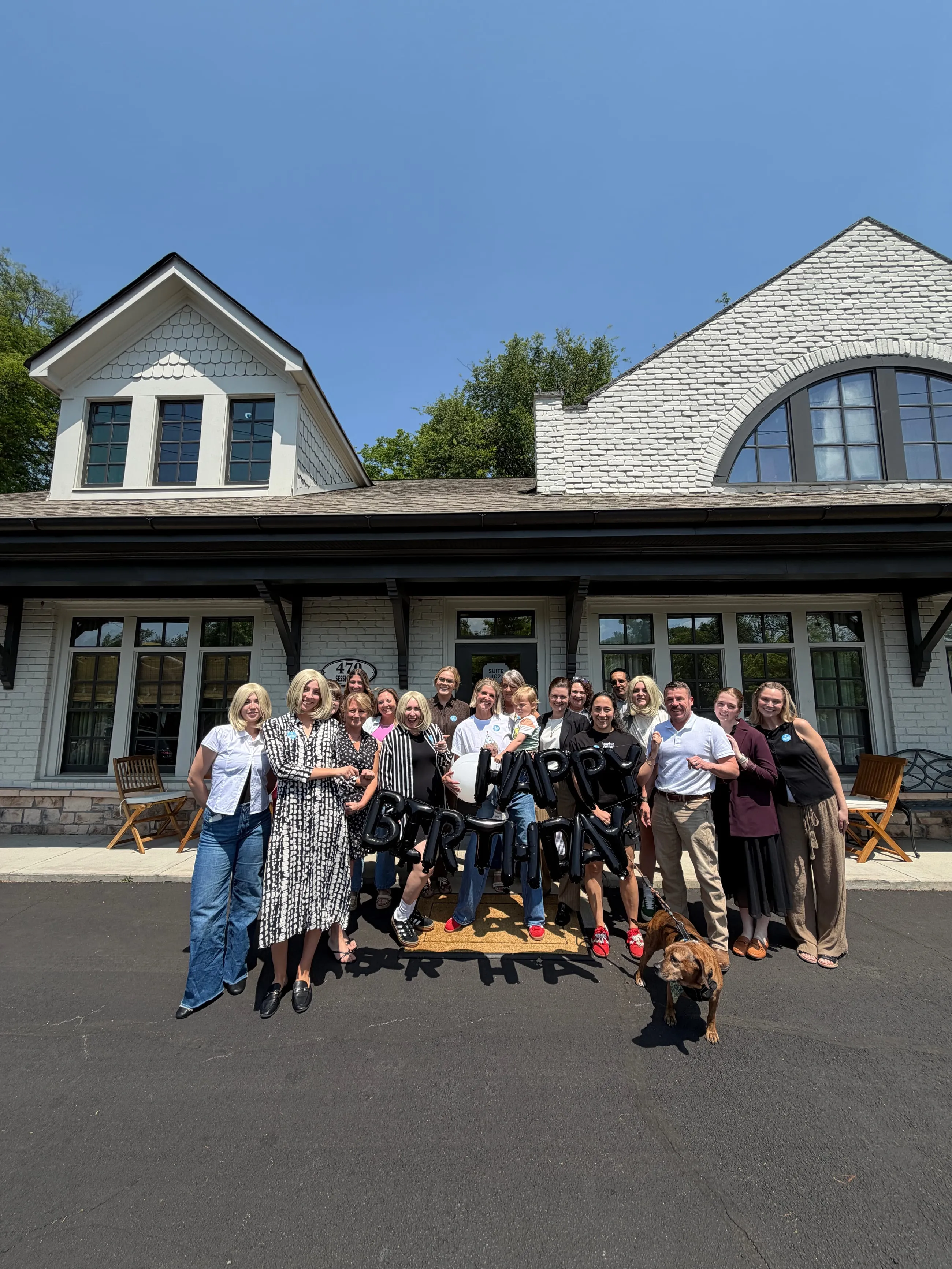 The team lined up on the drive in front of a white brick building with dark window frames, a dog at their feet, under a clear blue sky