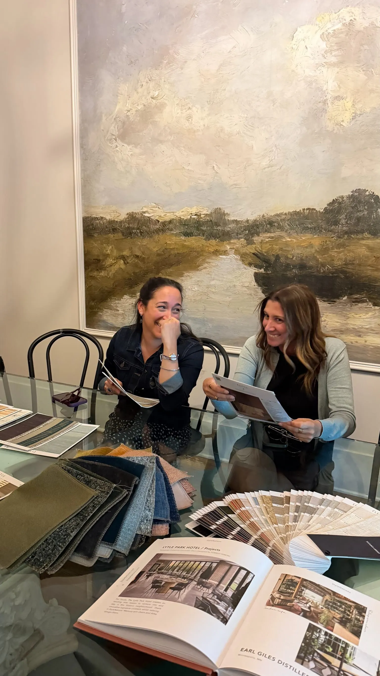 Two designers at a table leafing through a fabric sample book and an open catalogue beneath a landscape painting