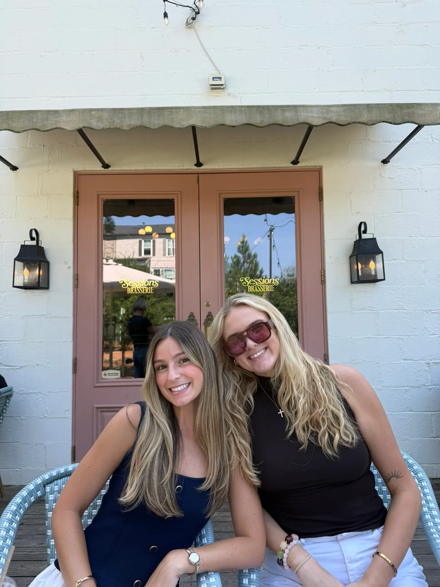 Two colleagues smiling cheek to cheek on a patio in front of the pink doors of Sessions Brasserie, one in sunglasses
