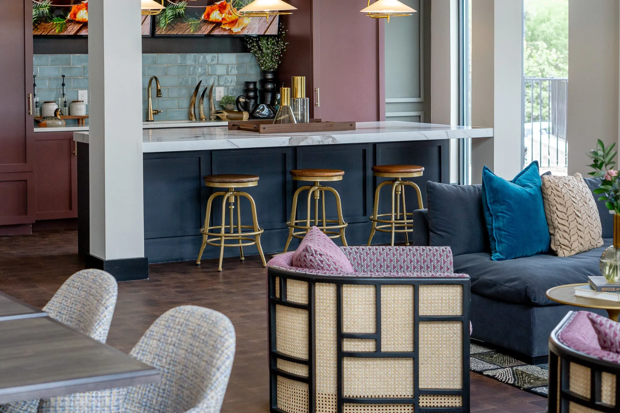 Clubroom kitchen with a navy island and brass stools, a teal velvet sofa and caned lounge chairs in the foreground