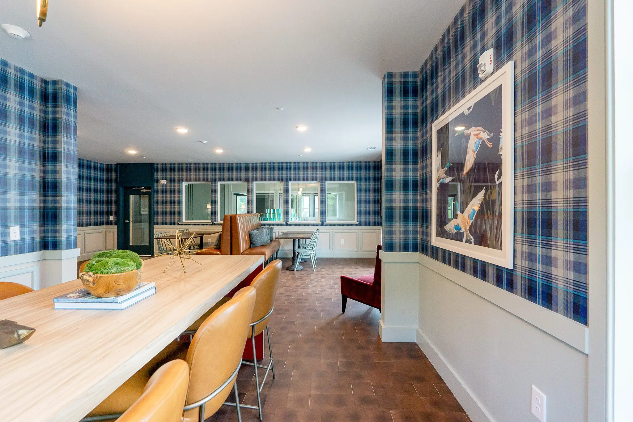 Clubroom with navy plaid wallpaper, a framed bird print and tan leather stools along a long pale wood table