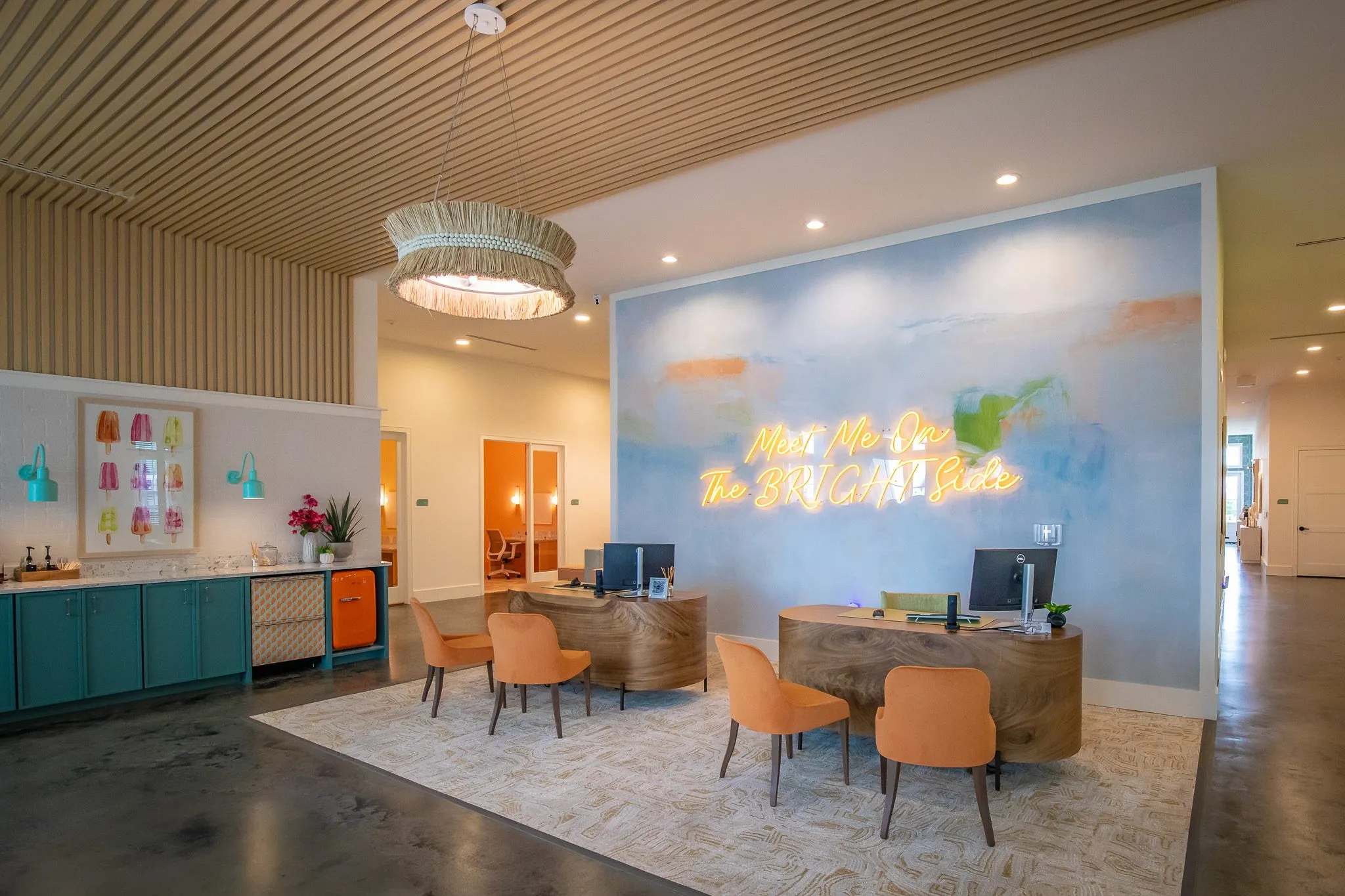 Leasing office with a slatted wood ceiling, coral chairs at two wood desks and a neon script sign on a sky blue mural wall