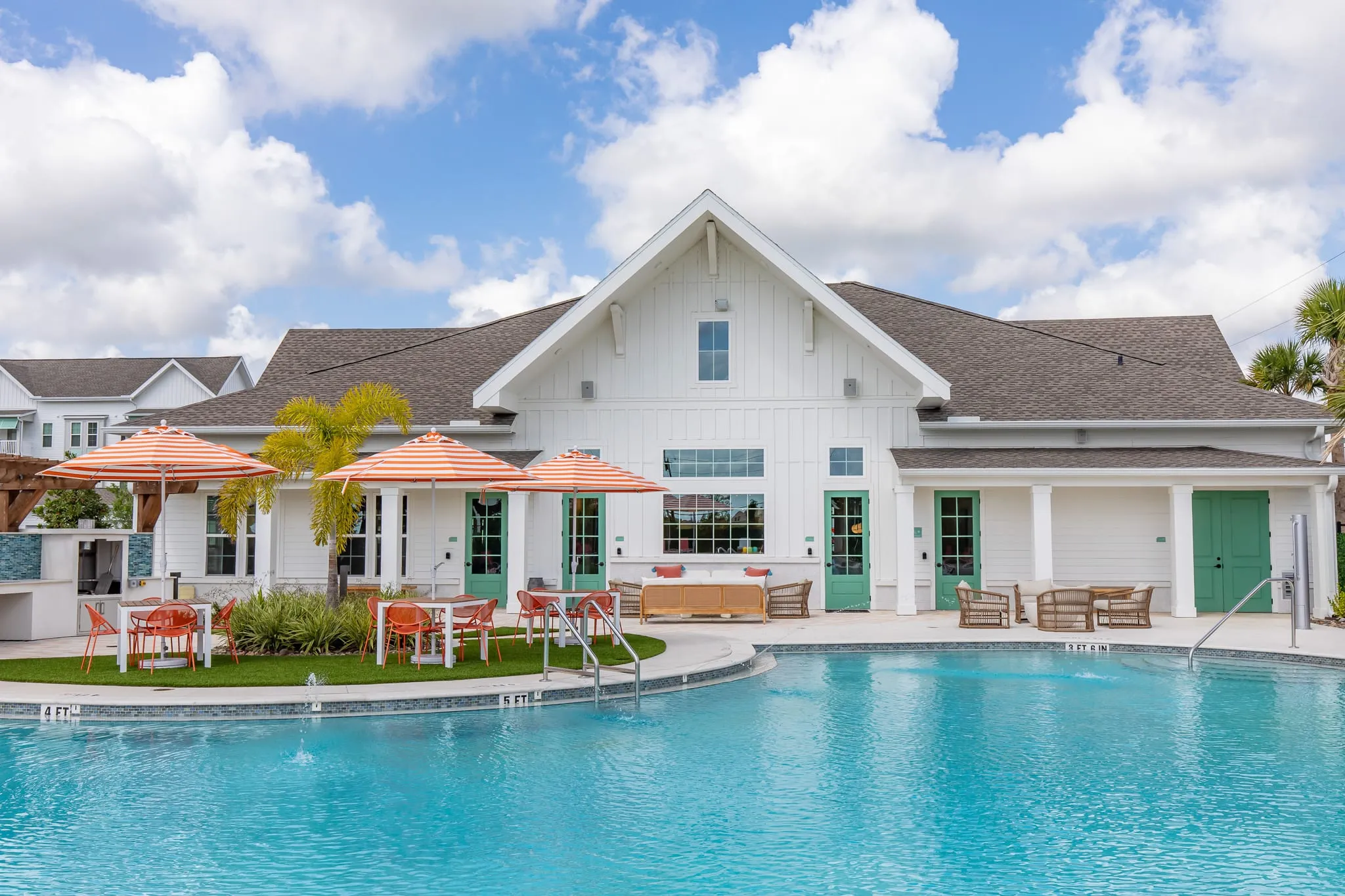 Multifamily community exterior: clubhouse with a white gabled roofline across the pool, coral umbrellas on the deck