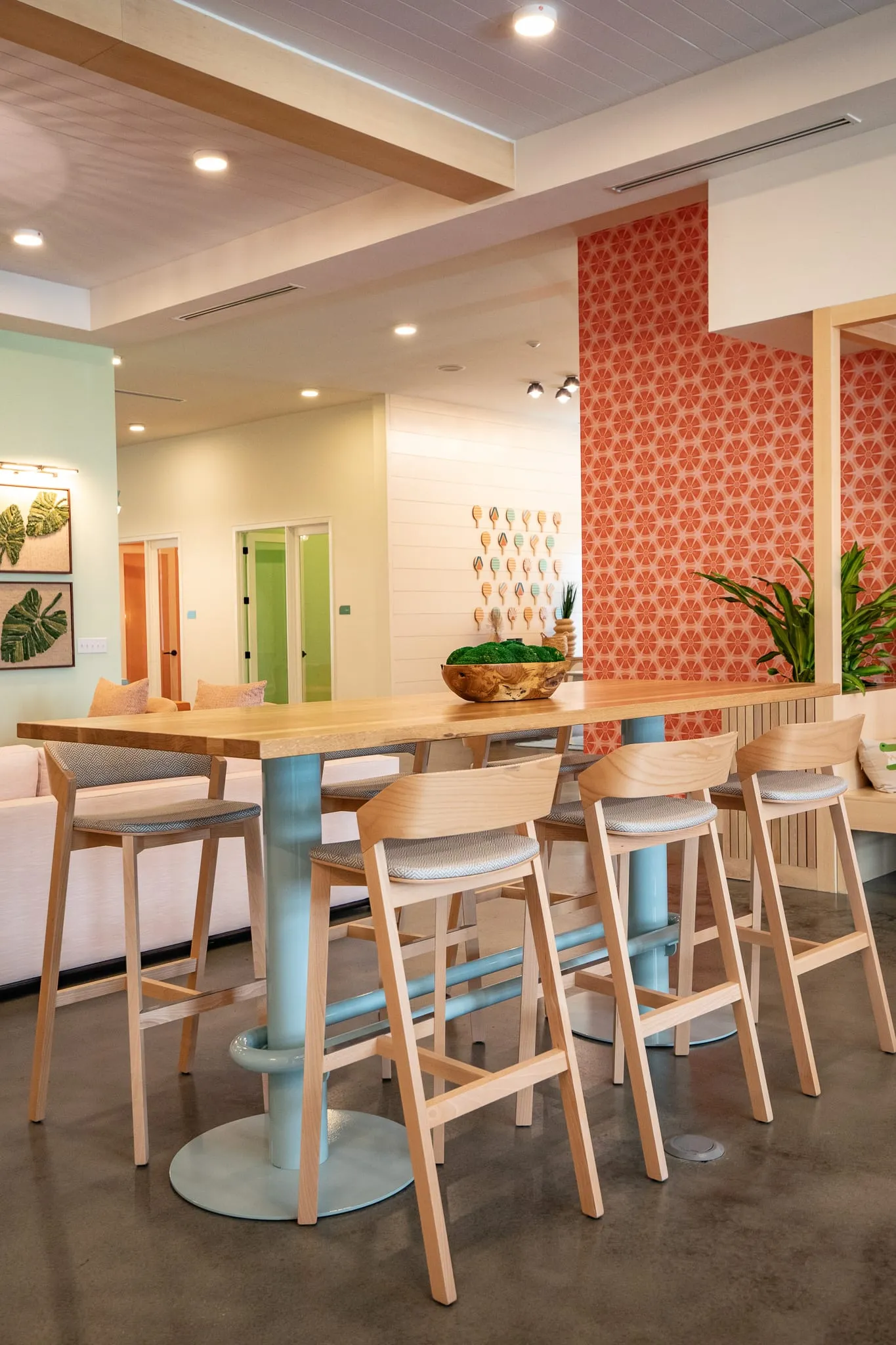 Multifamily amenity lounge with blond wood stools at a high-top table on a teal base before coral patterned wallpaper