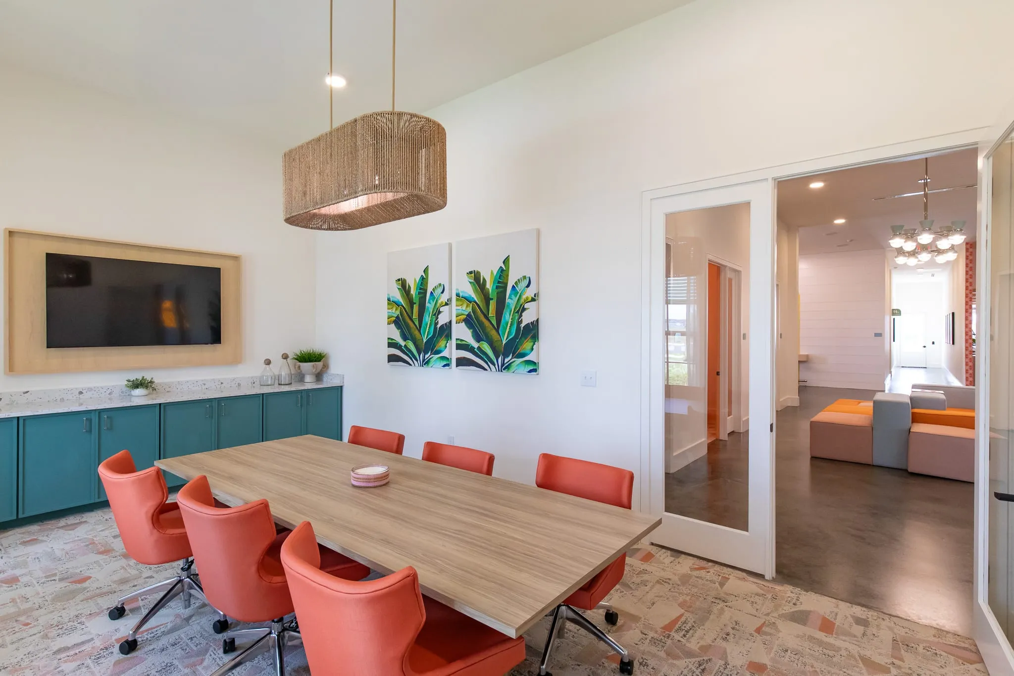 Multifamily amenity conference room with coral task chairs around a long wood table below banana leaf prints