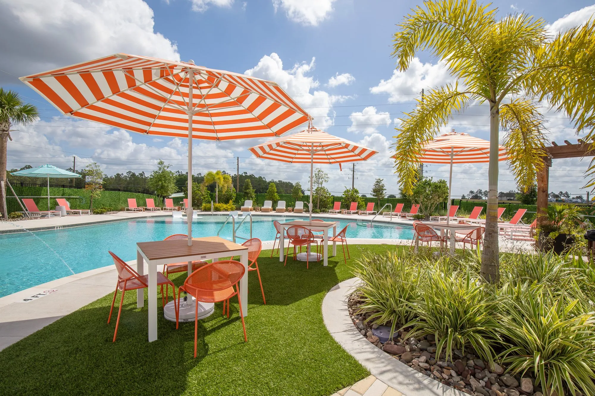 Multifamily amenity pool with striped umbrellas over tables and coral chairs on turf beside a palm and planting