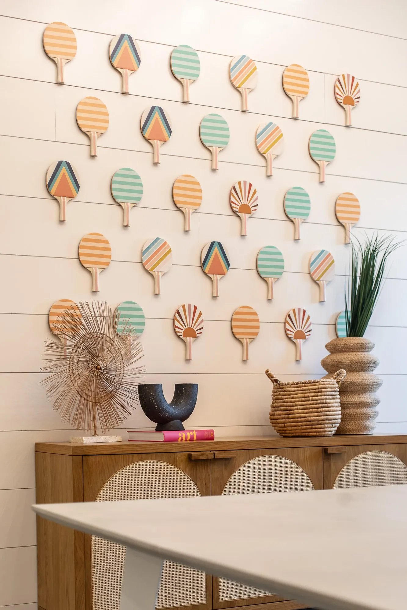 Multifamily interior detail: painted ping pong paddles on white shiplap above a cane front console with baskets and vases