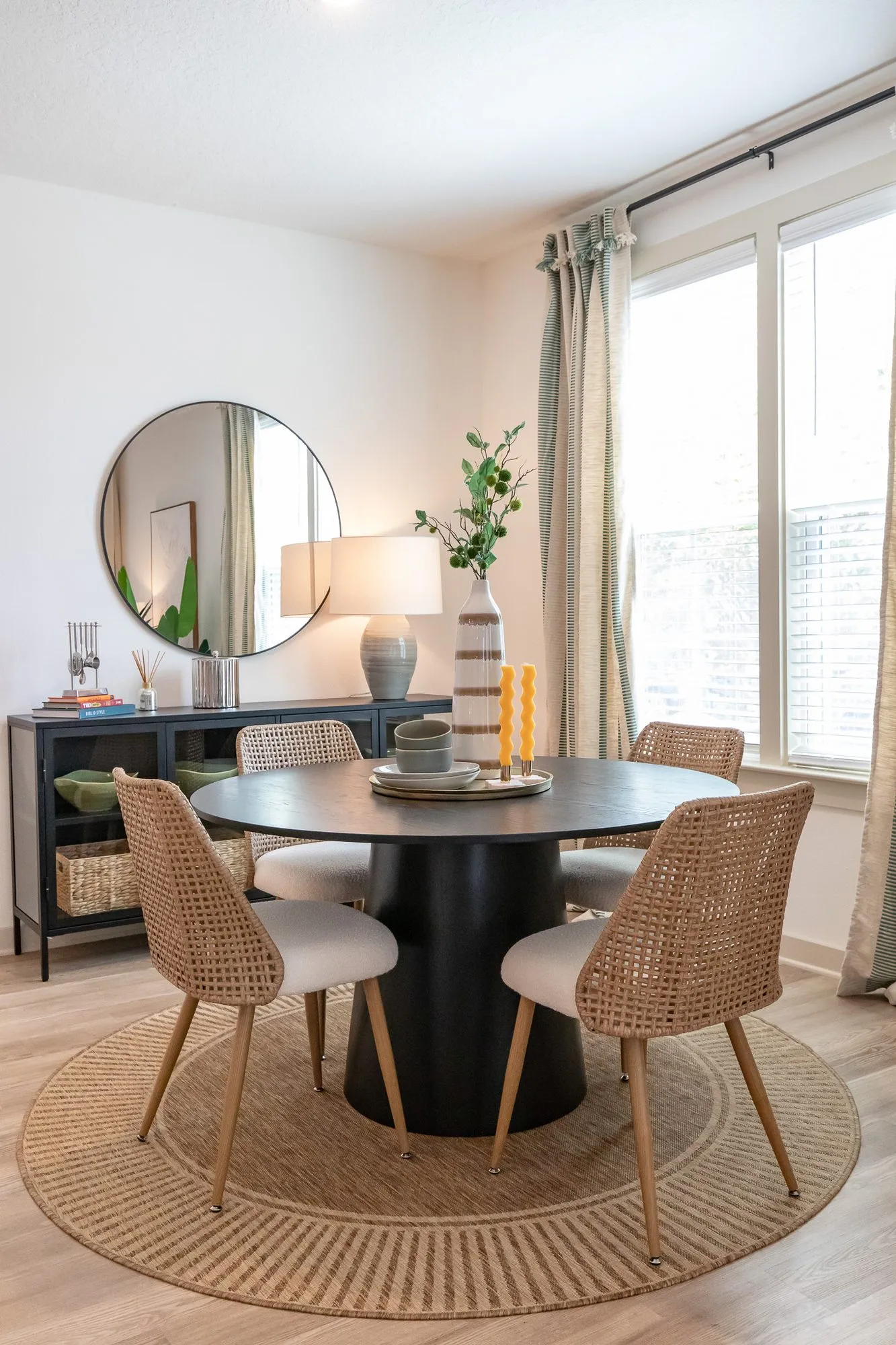 Multifamily model apartment dining nook with a round black table and cane chairs on a jute rug beside tall windows