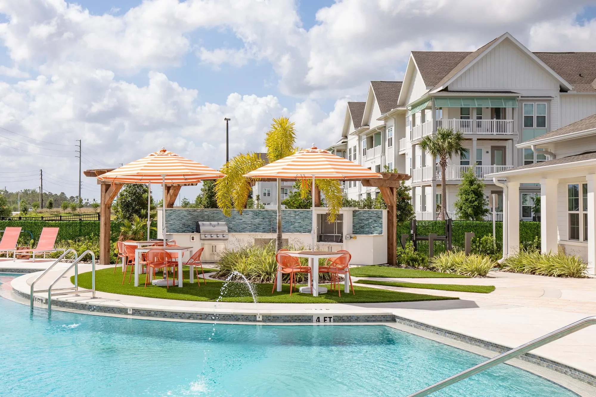 Multifamily amenity pool with striped umbrellas over tables and an outdoor kitchen under a timber pergola, apartments behind