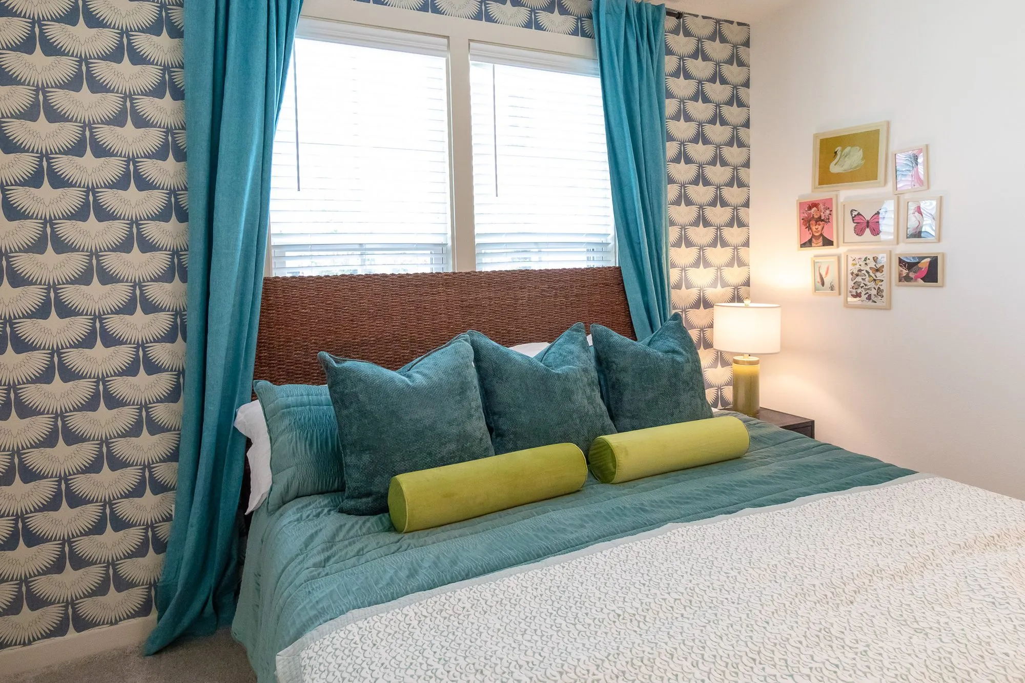 Multifamily apartment bedroom with blue crane wallpaper, teal curtains, a woven headboard and a gallery of small prints
