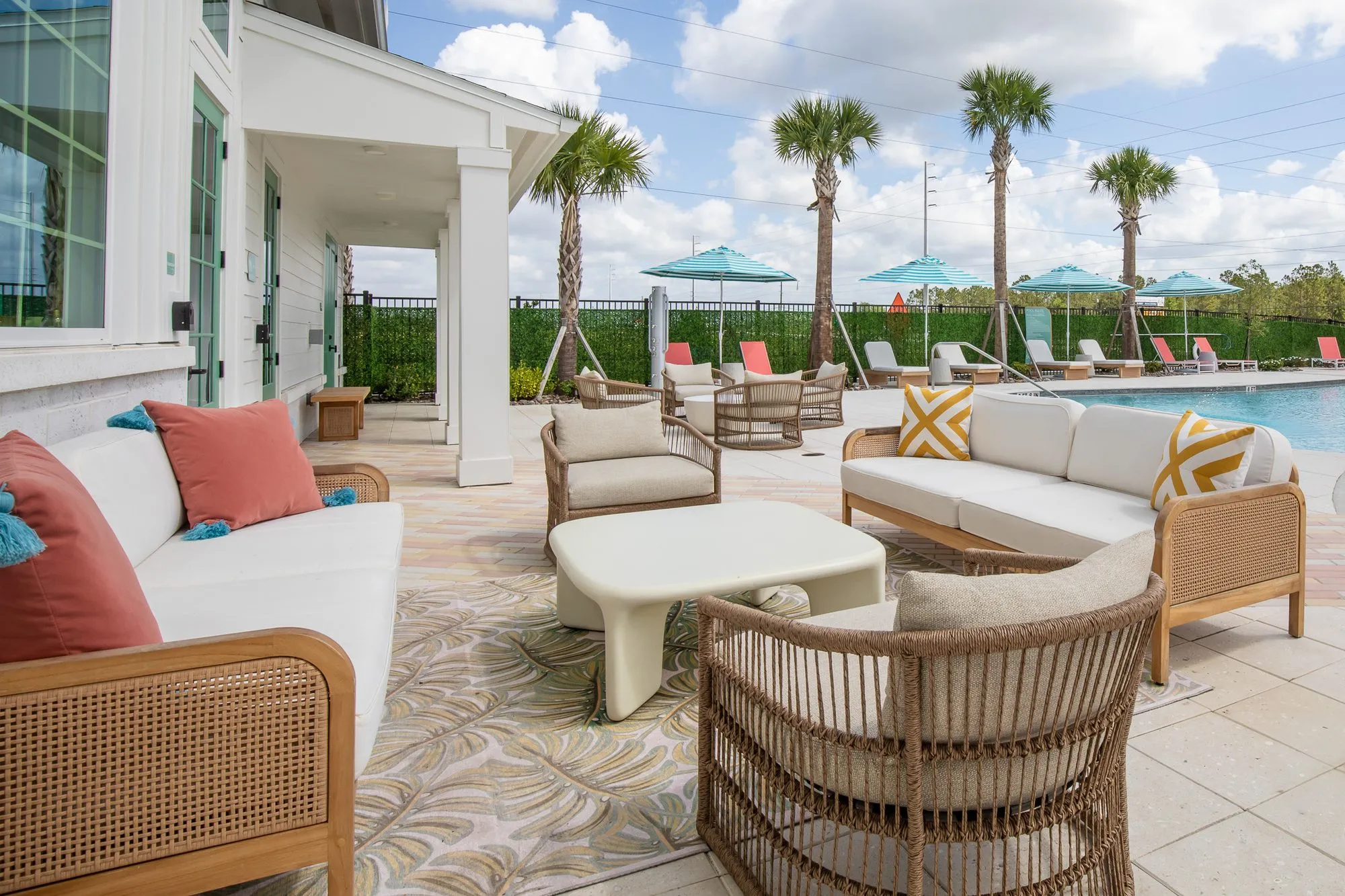 Multifamily amenity covered patio with a rattan sofa and chairs, coral and yellow pillows, pool and palms beyond