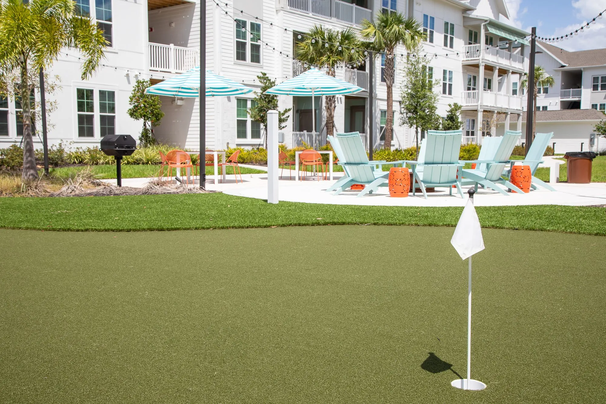 Multifamily amenity putting green with a flag, blue Adirondack chairs and umbrellas on the courtyard beyond