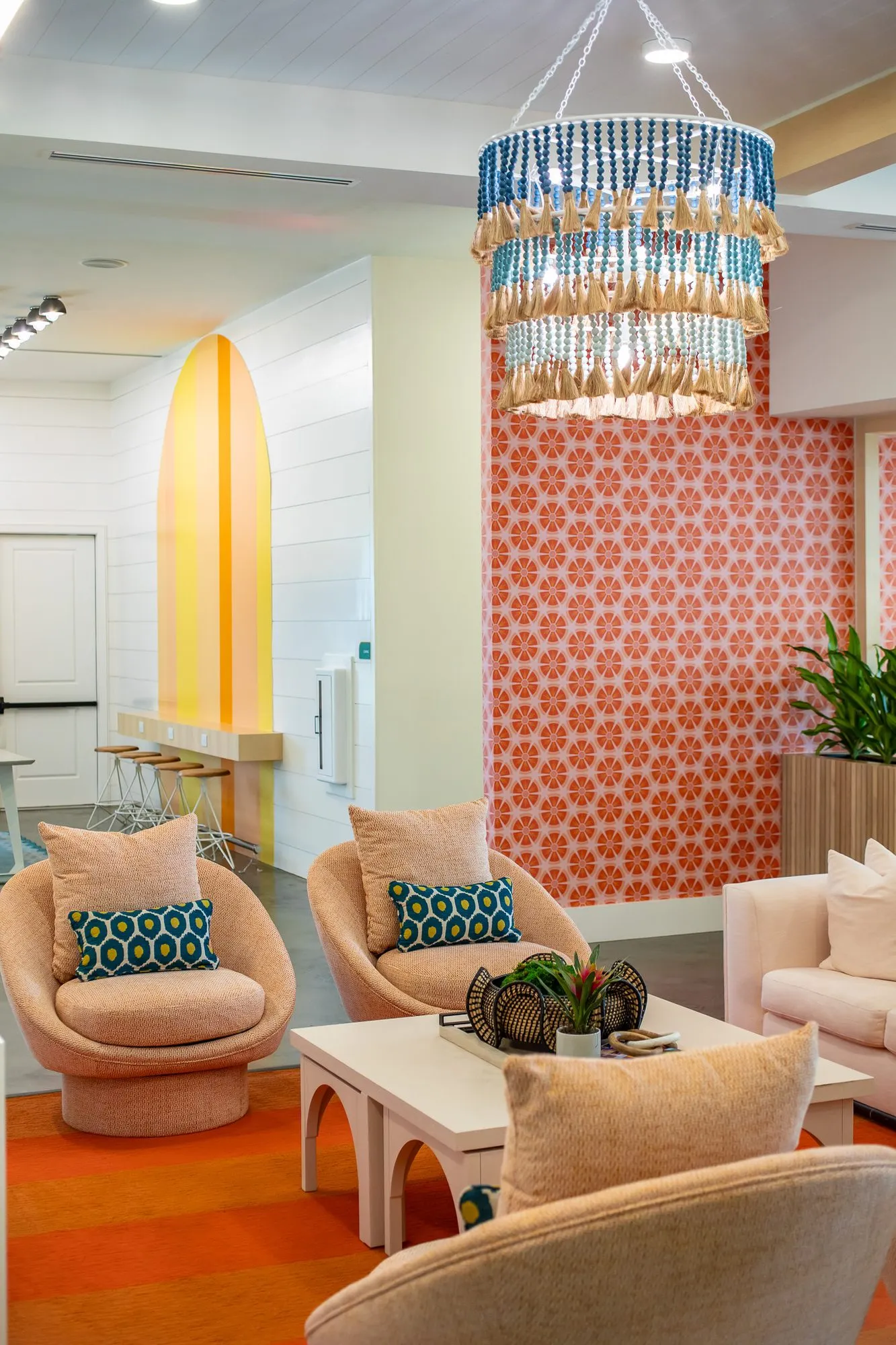 Multifamily amenity lounge with rattan swivel chairs under a beaded chandelier, a yellow arched niche and coral wallpaper
