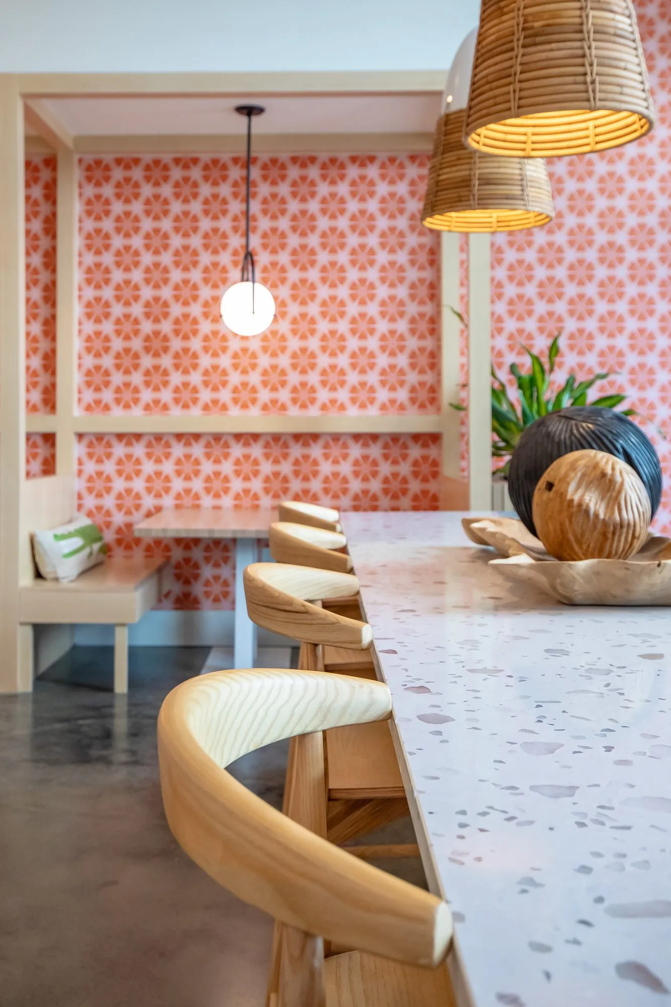 Multifamily amenity terrazzo bar with blond wood stools under rattan pendants, coral patterned booth seating beyond