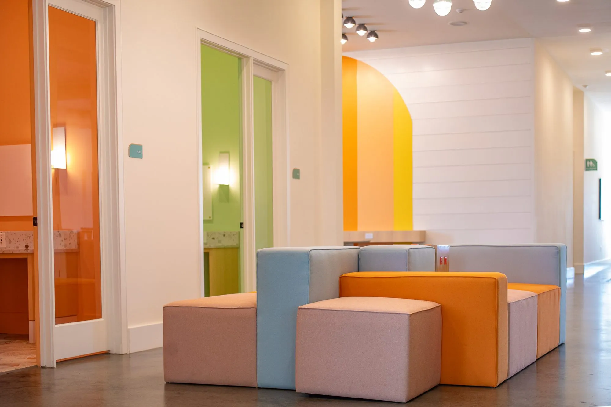 Multifamily amenity corridor with modular cube seating, glass doors to orange and green offices and a striped arch beyond