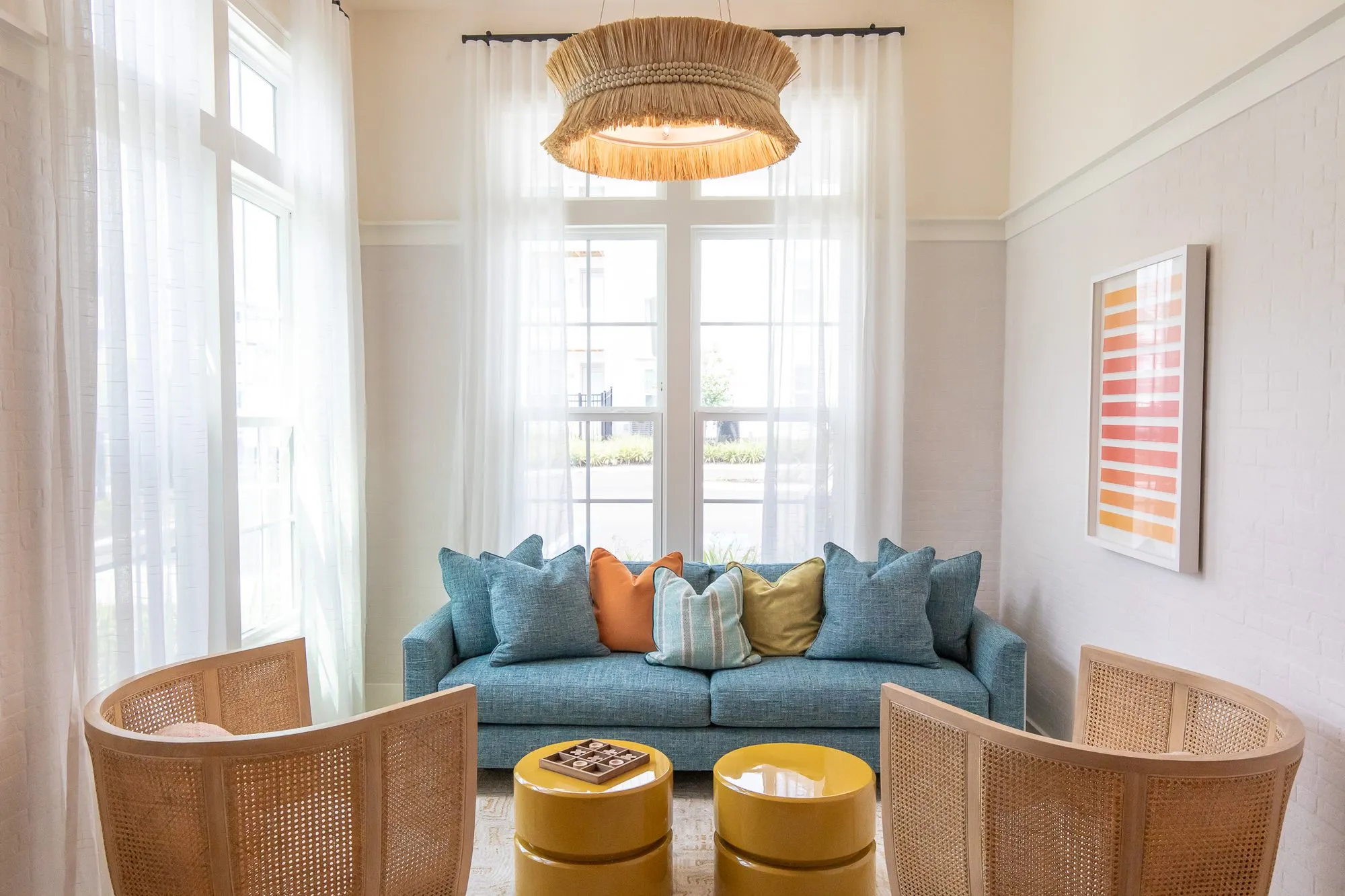 Multifamily sitting room with cane barrel chairs and yellow drum tables before a blue sofa, striped art at right