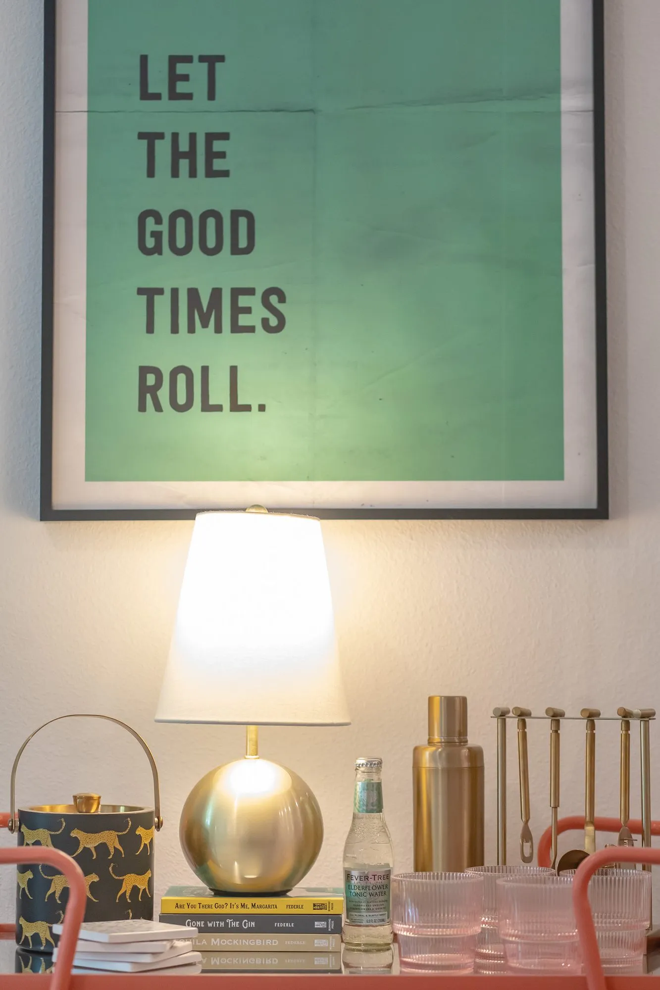 Multifamily interior detail: bar cart with a brass lamp, glassware and ice bucket below a green Let the Good Times Roll print