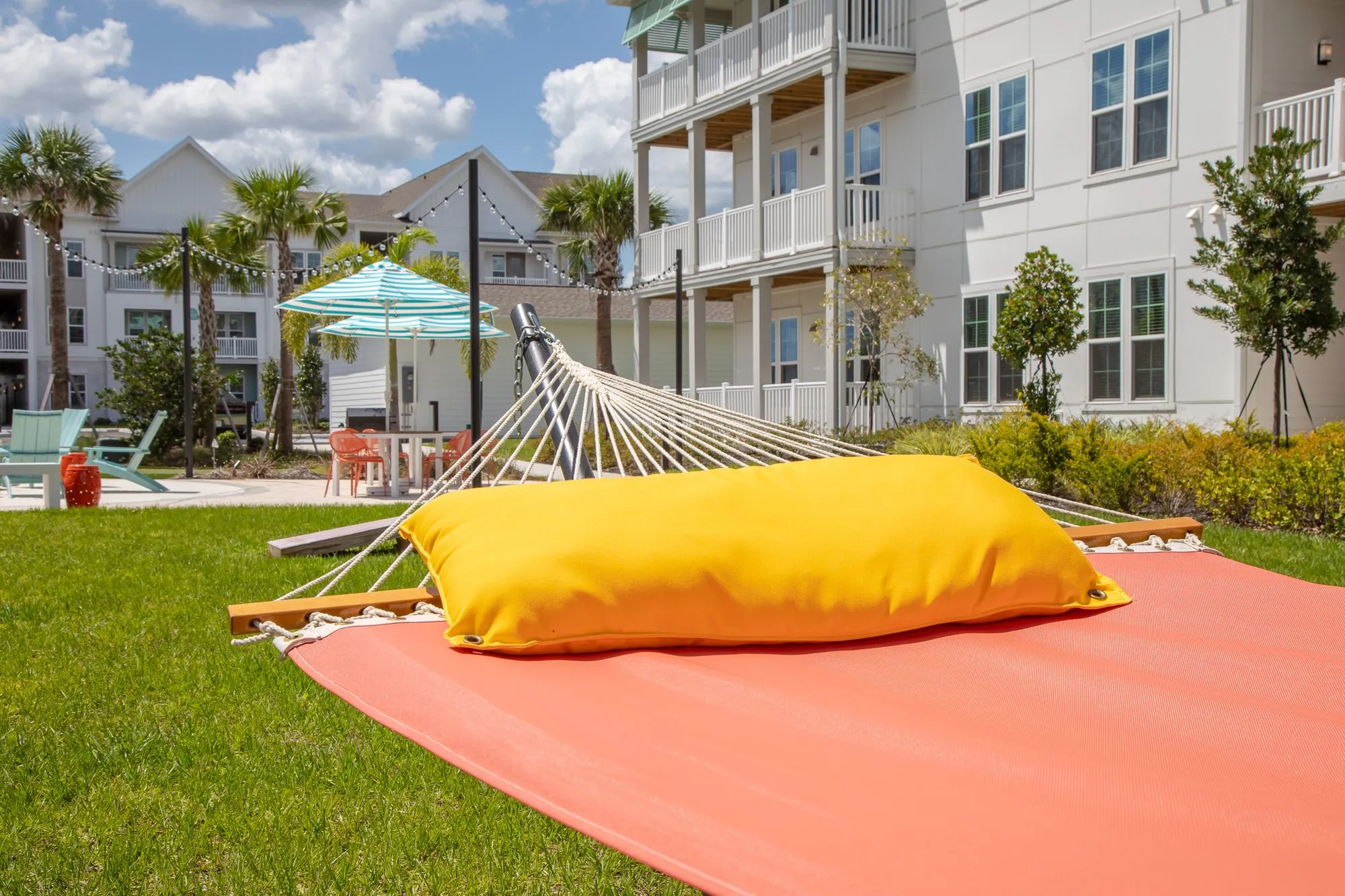 Multifamily amenity lawn detail: coral hammock with a yellow pillow, umbrellas and apartment buildings behind