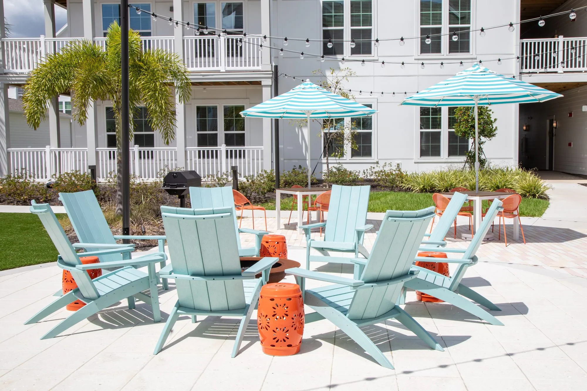 Multifamily amenity fire pit with pale blue Adirondack chairs before teal striped umbrellas, string lights overhead