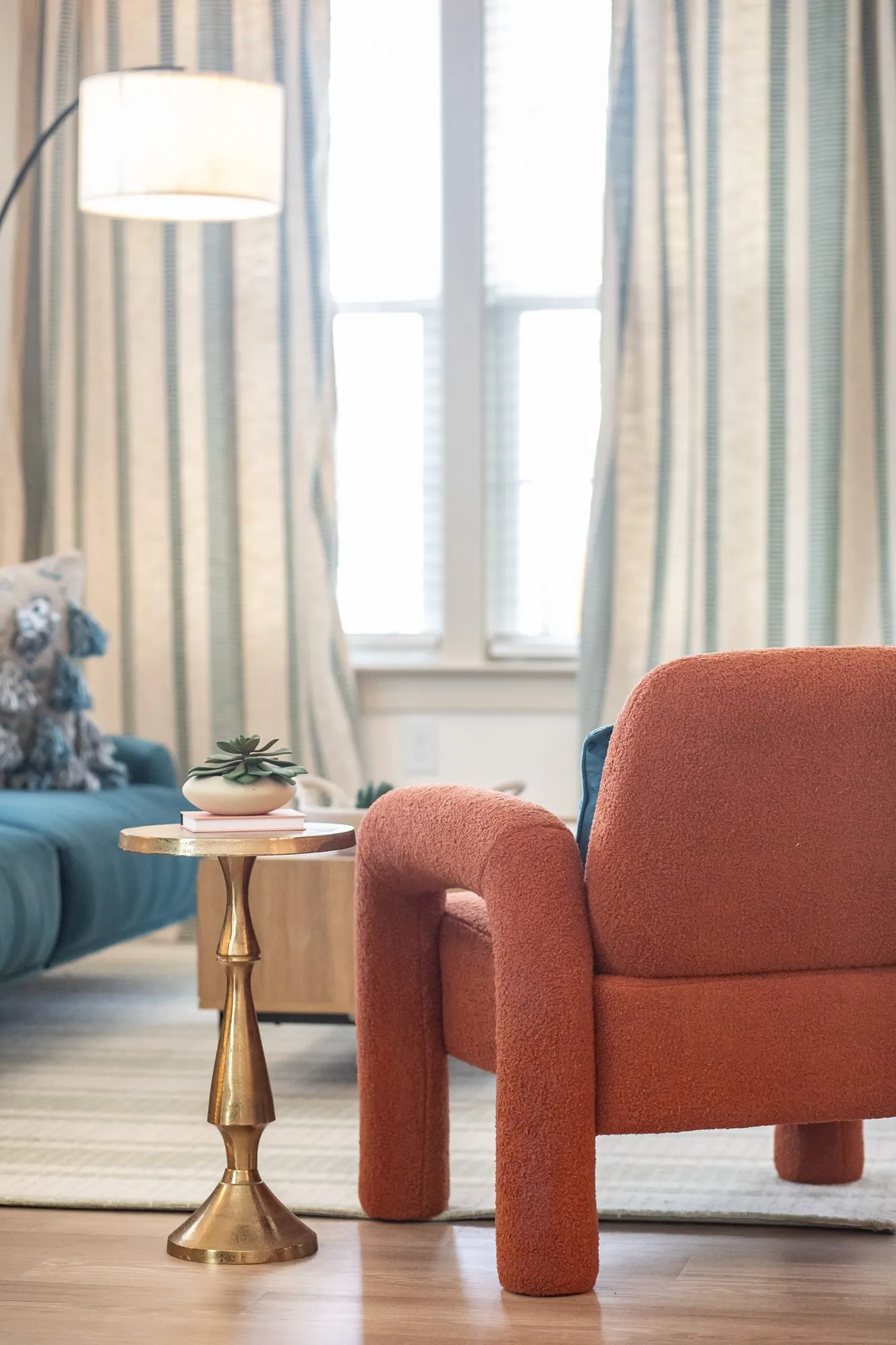 Multifamily interior detail: rust boucle armchair beside a brass pedestal side table and an arc floor lamp by curtains