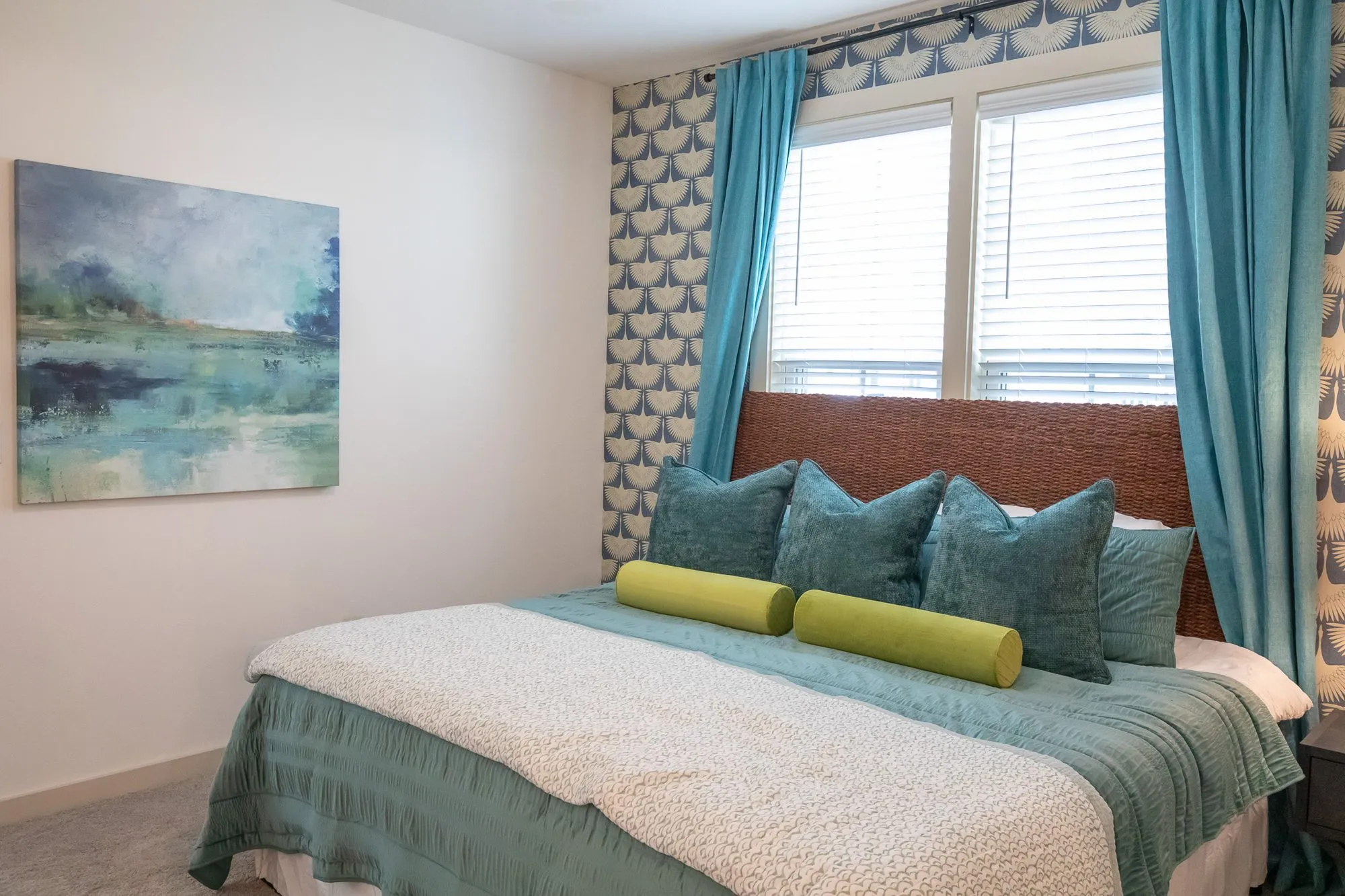 Multifamily apartment bedroom from the foot of the bed, teal pillows and yellow bolsters, a landscape painting, teal curtains