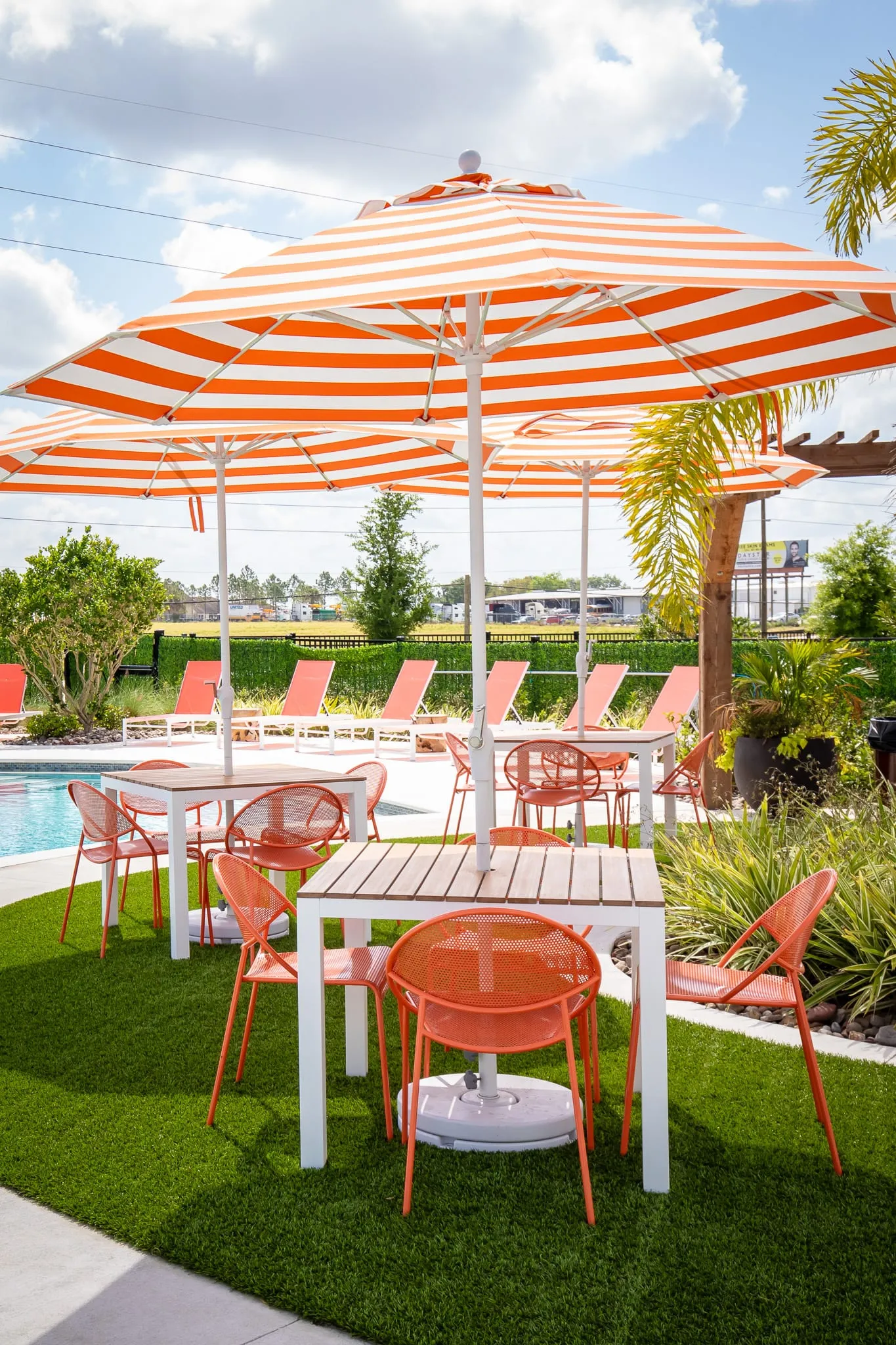 Multifamily amenity poolside dining with orange and white striped umbrellas over coral bistro chairs on turf