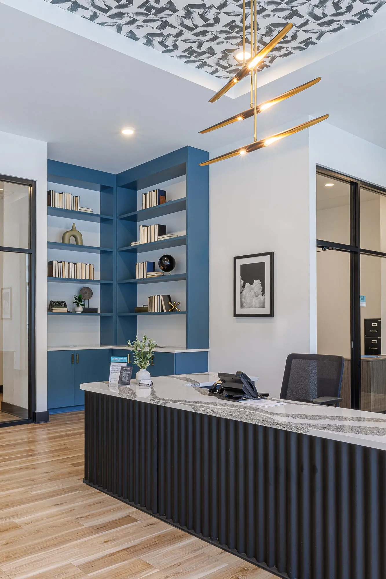 Multifamily amenity reception desk, fluted black with a marble top, before blue shelving under a brass stick chandelier