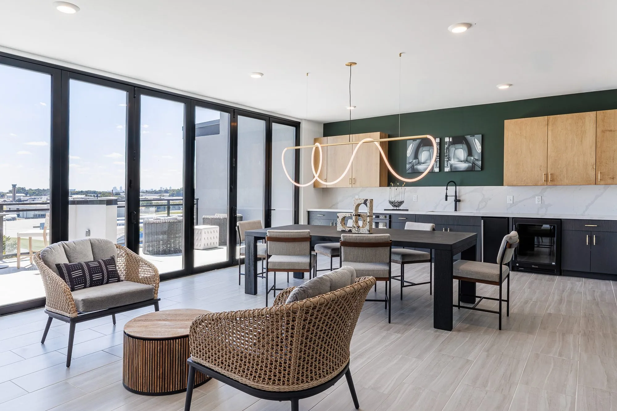 Multifamily amenity sky lounge with woven armchairs by folding glass walls and a dining table under a looping rope light