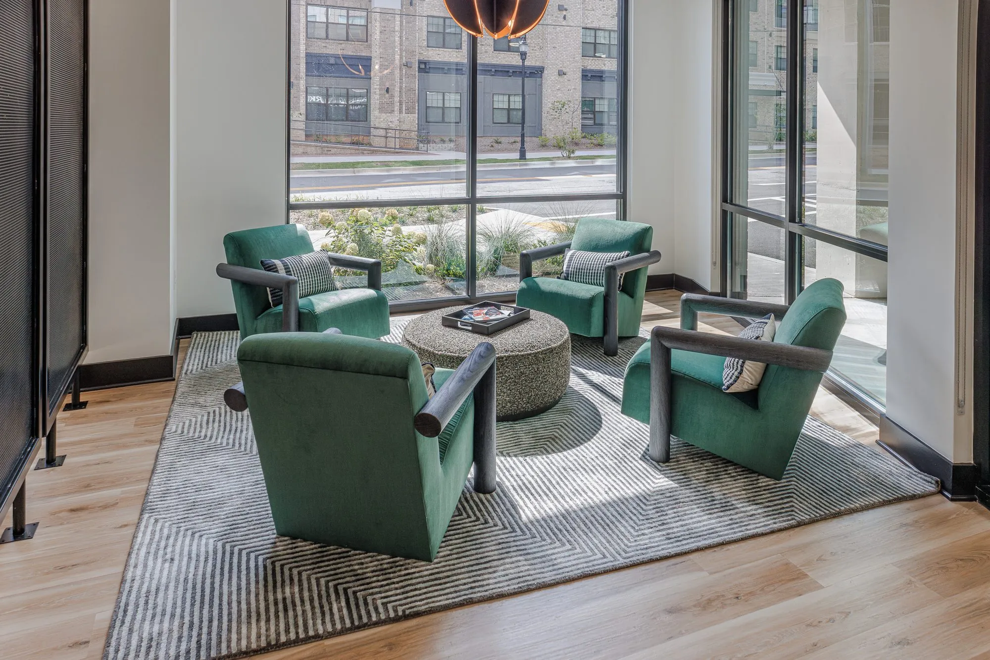 Multifamily interior detail: four green velvet armchairs around a round ottoman on a striped rug in a glazed corner