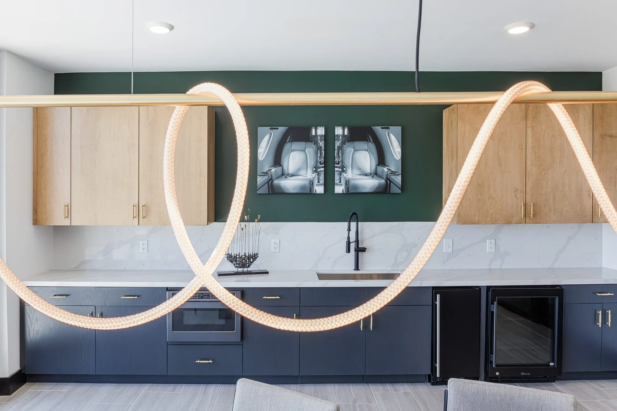 Multifamily amenity kitchenette with navy and oak cabinetry and aircraft cabin photographs on green, a rope light in front